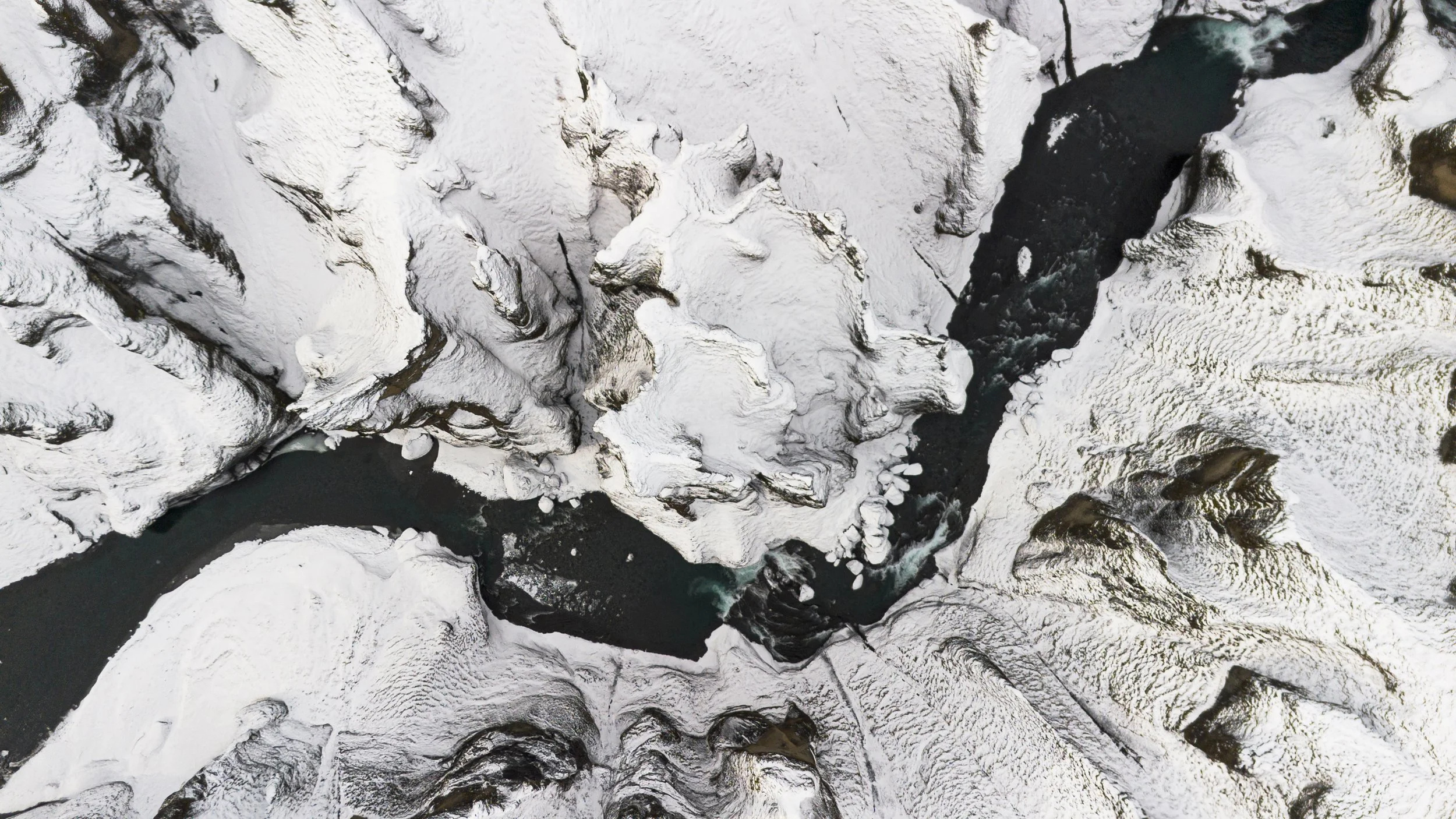 Aerial view of a snowy canyon with a dark river flowing through it, surrounded by ice-covered cliffs.