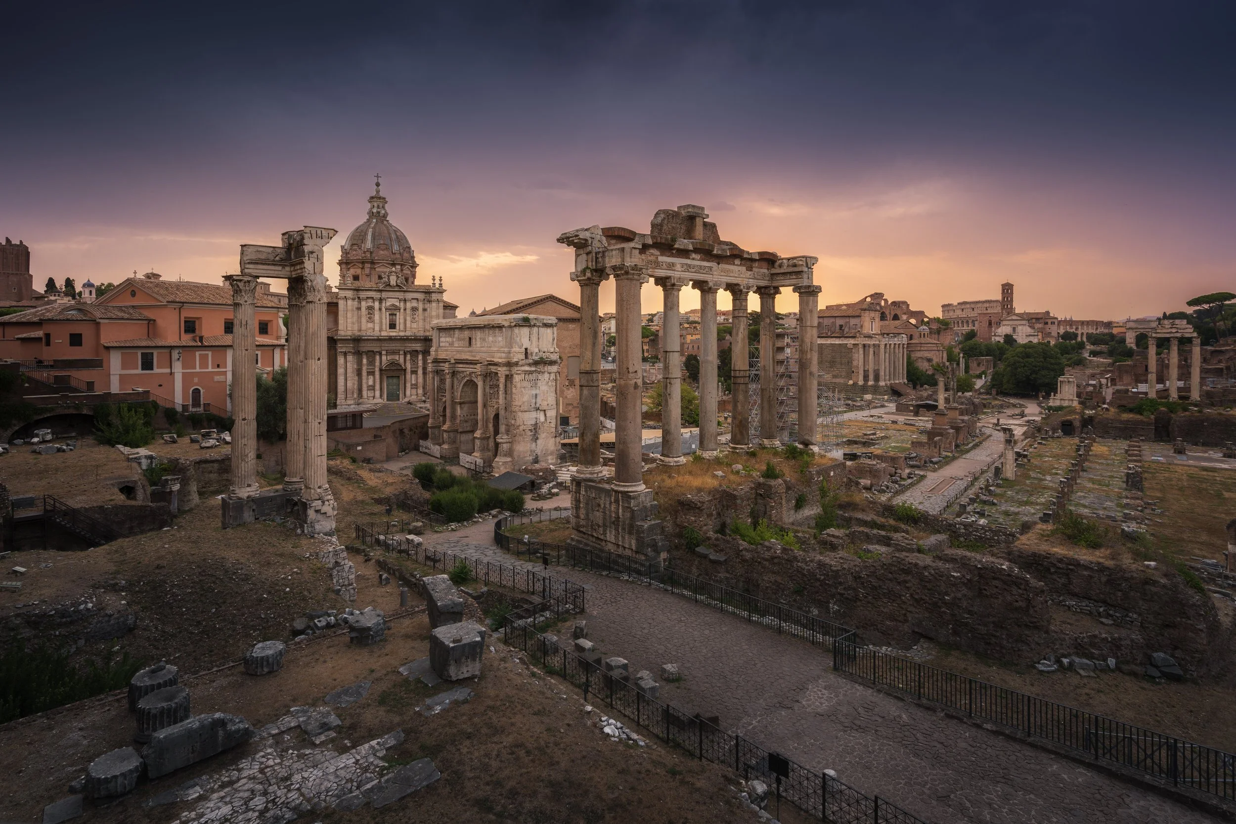 Ancient ruins of Roman temples and structures at sunset, with cloudy sky and cityscape in the background.