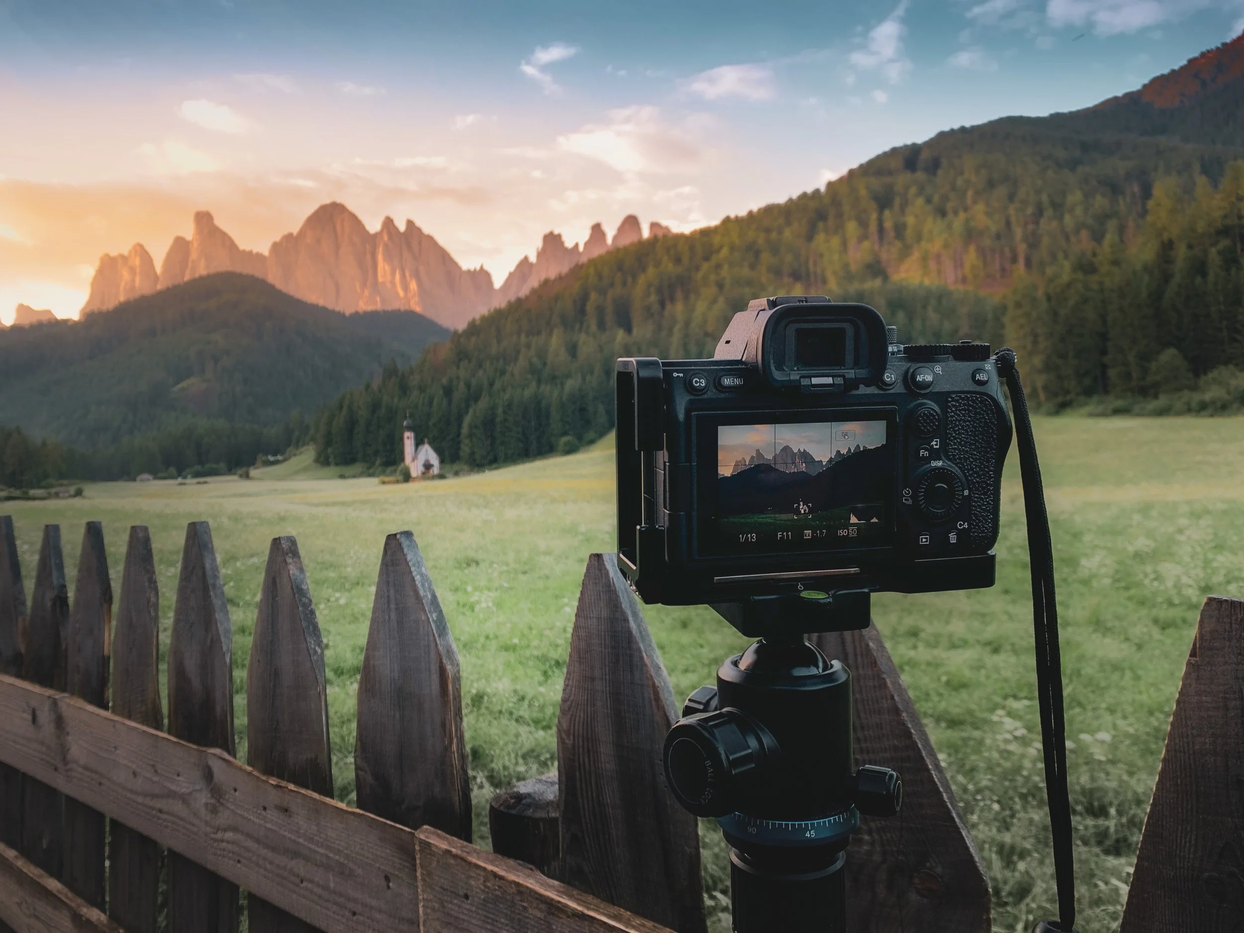 A camera on a tripod capturing a landscape scene of mountains, a church, and a green field during sunset.