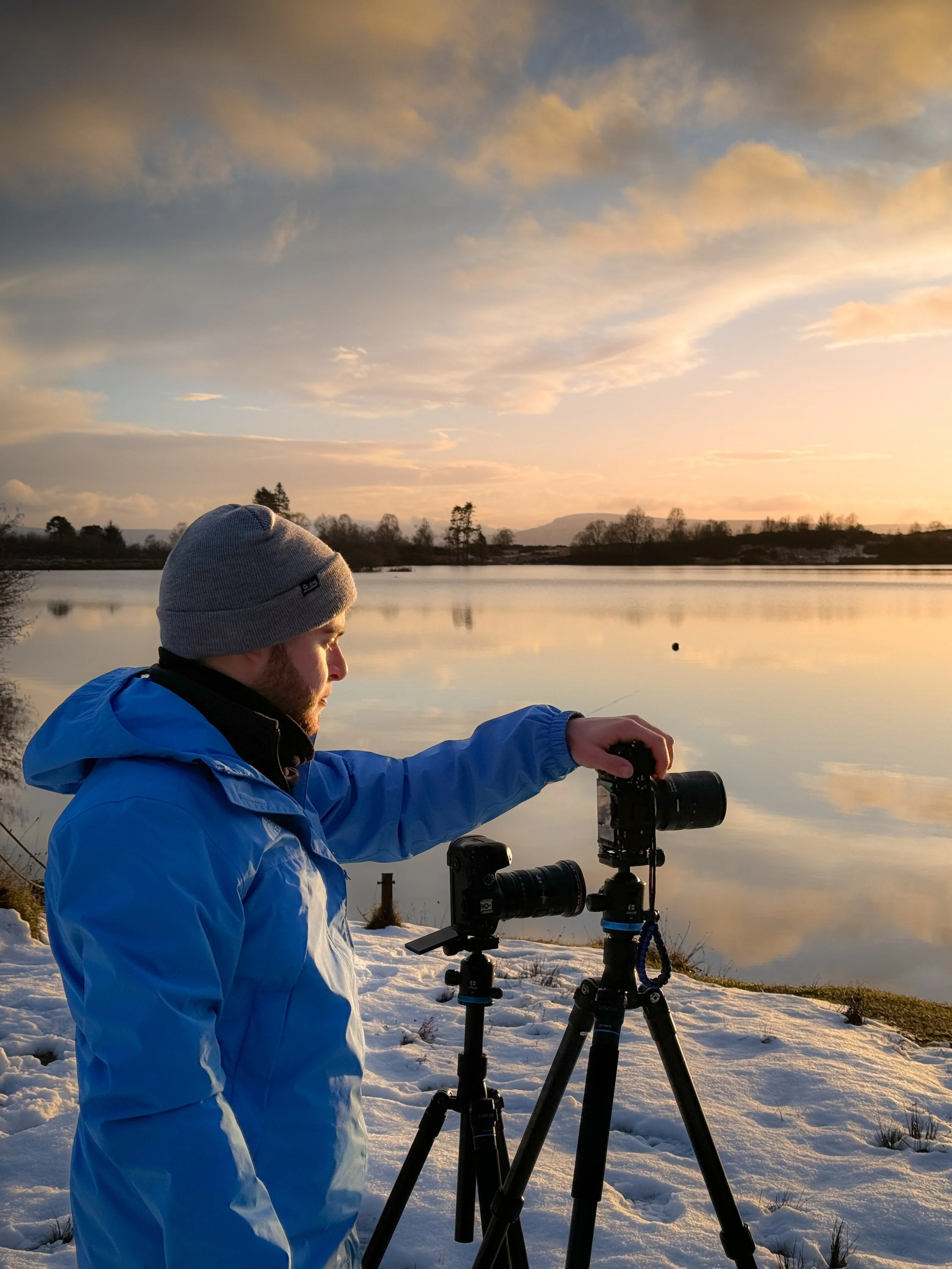 Man in a blue jacket and gray beanie setting up cameras on tripods by a lake at sunset with snow on the ground.