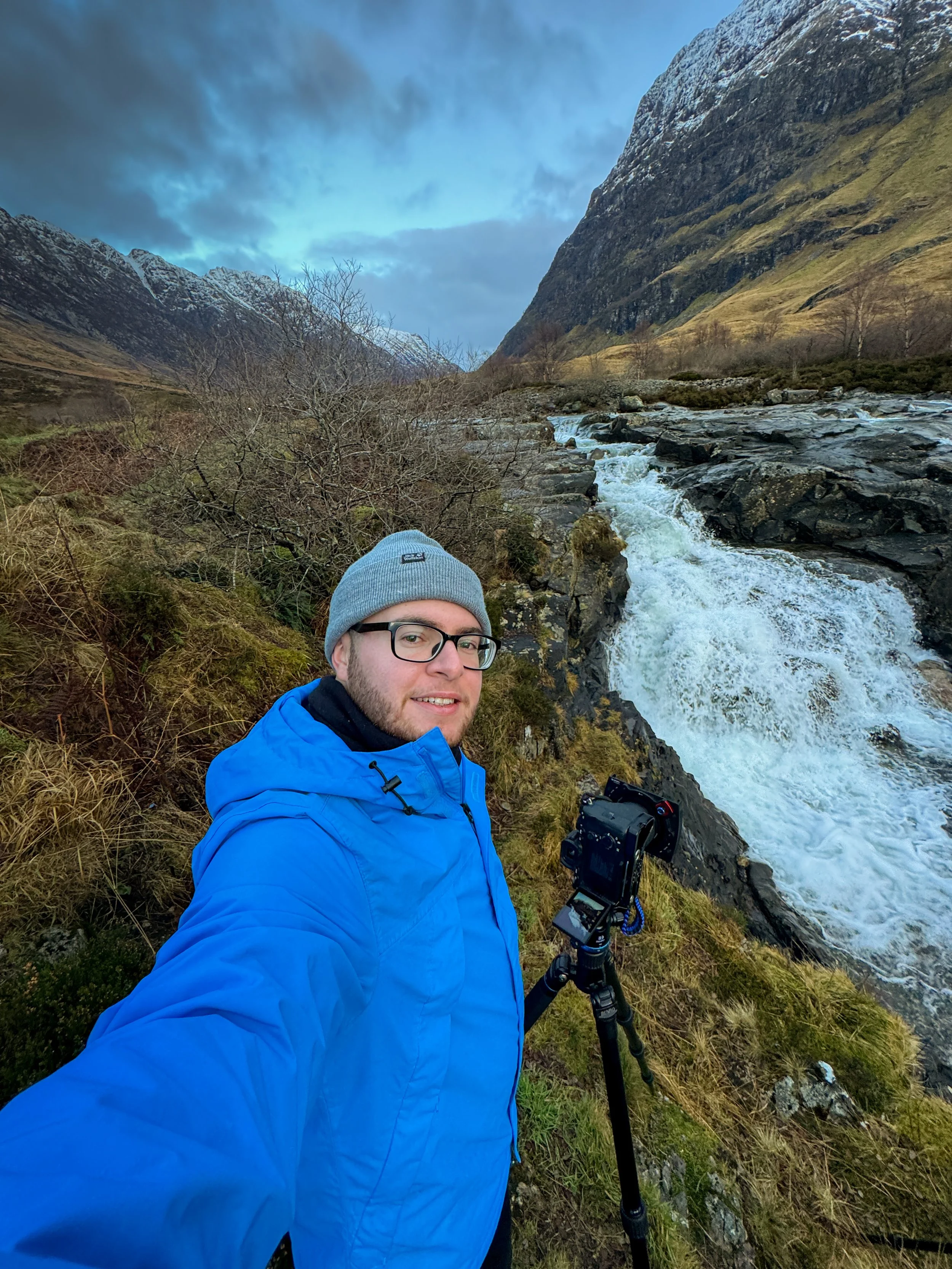 A man wearing glasses, a gray beanie, and a bright blue jacket takes a selfie near a flowing river in a mountainous landscape with snow-capped peaks and cloudy sky.