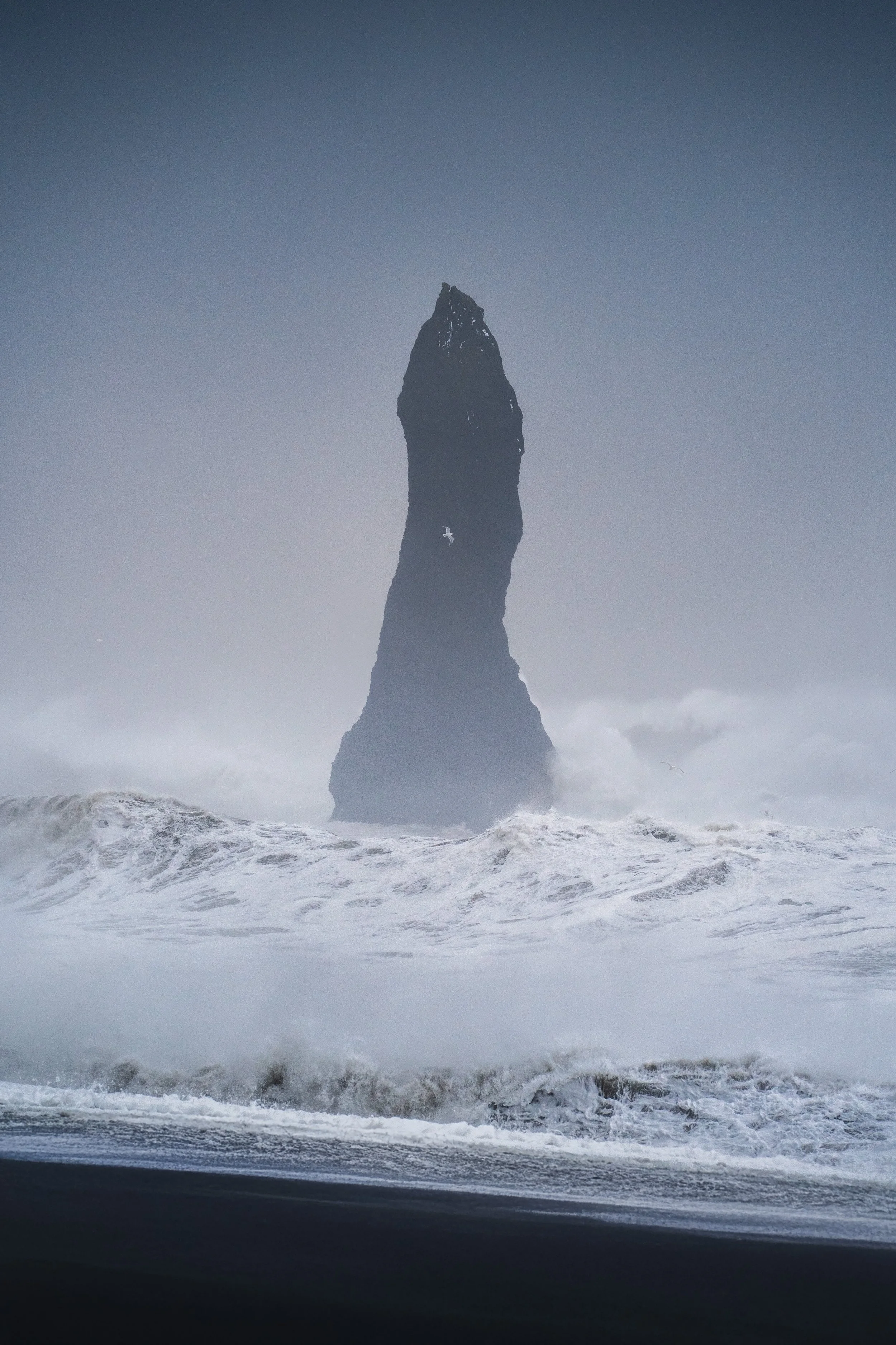 A tall, dark sea stack rising from choppy ocean waters with waves crashing around its base, under a cloudy, overcast sky.
