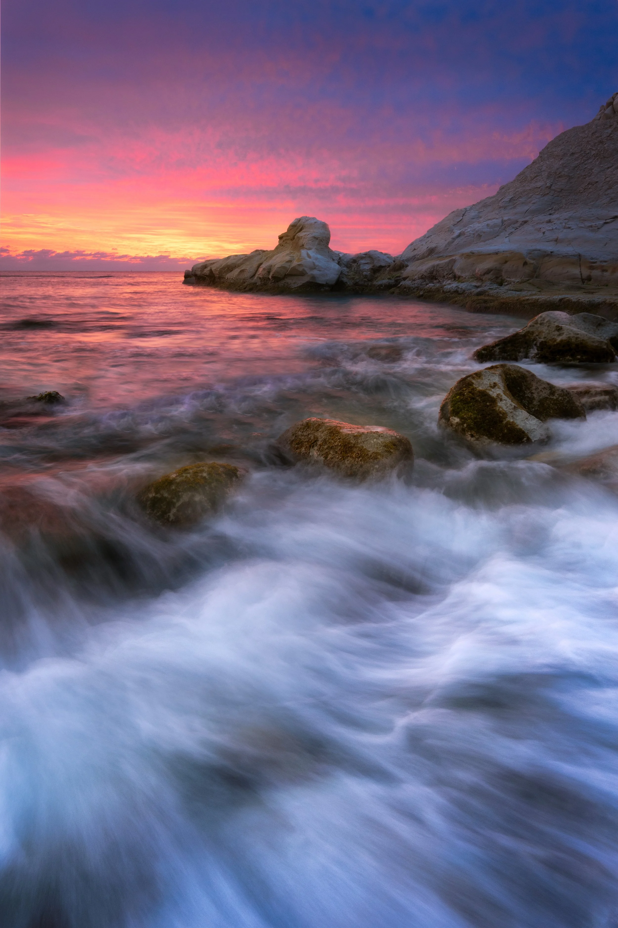 Colorful sunset over the ocean with rocks and gentle waves in the foreground.