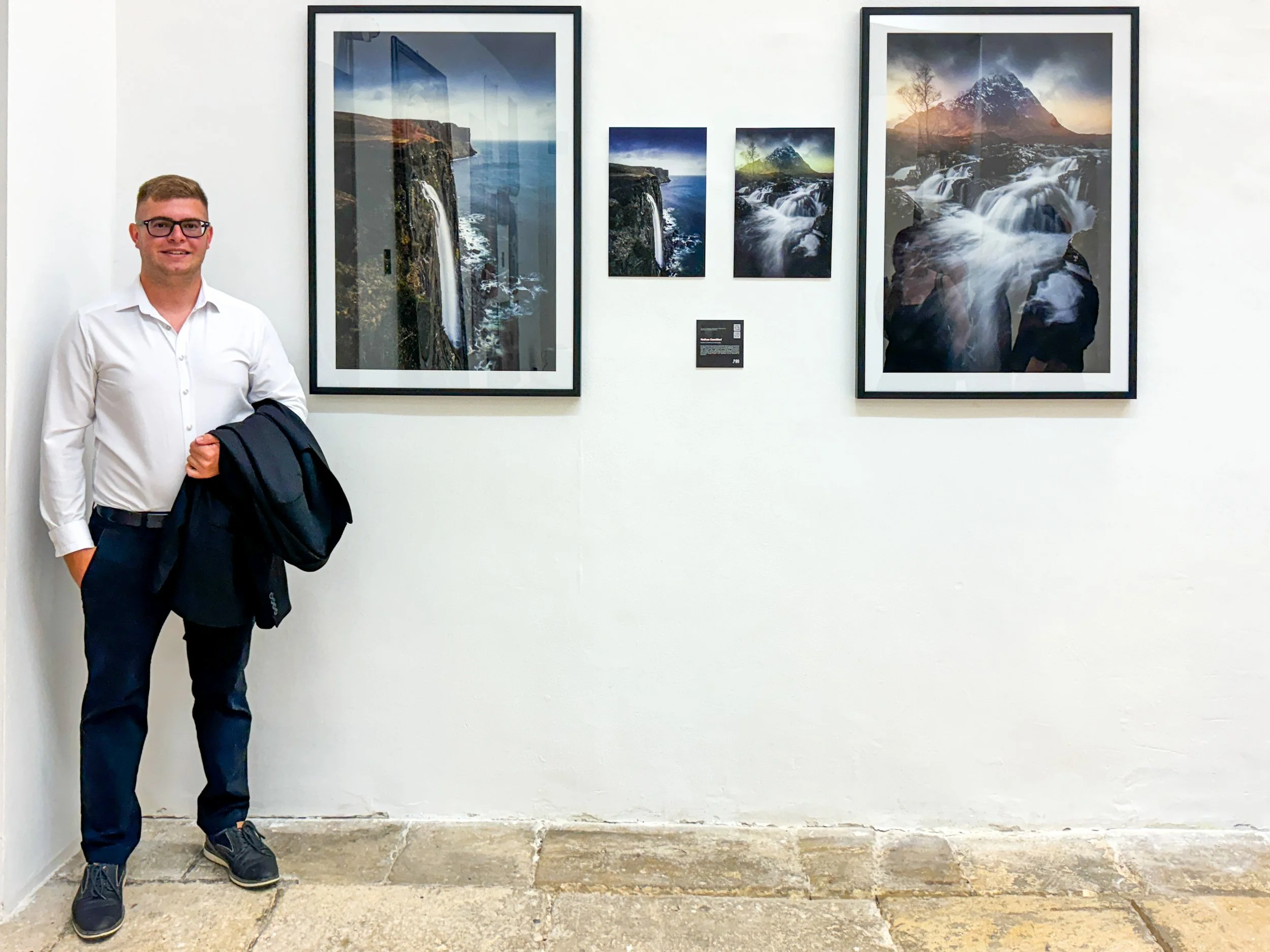 A man in a white shirt holding a dark jacket stands next to a white wall displaying five framed landscape photographs of waterfalls and mountains.