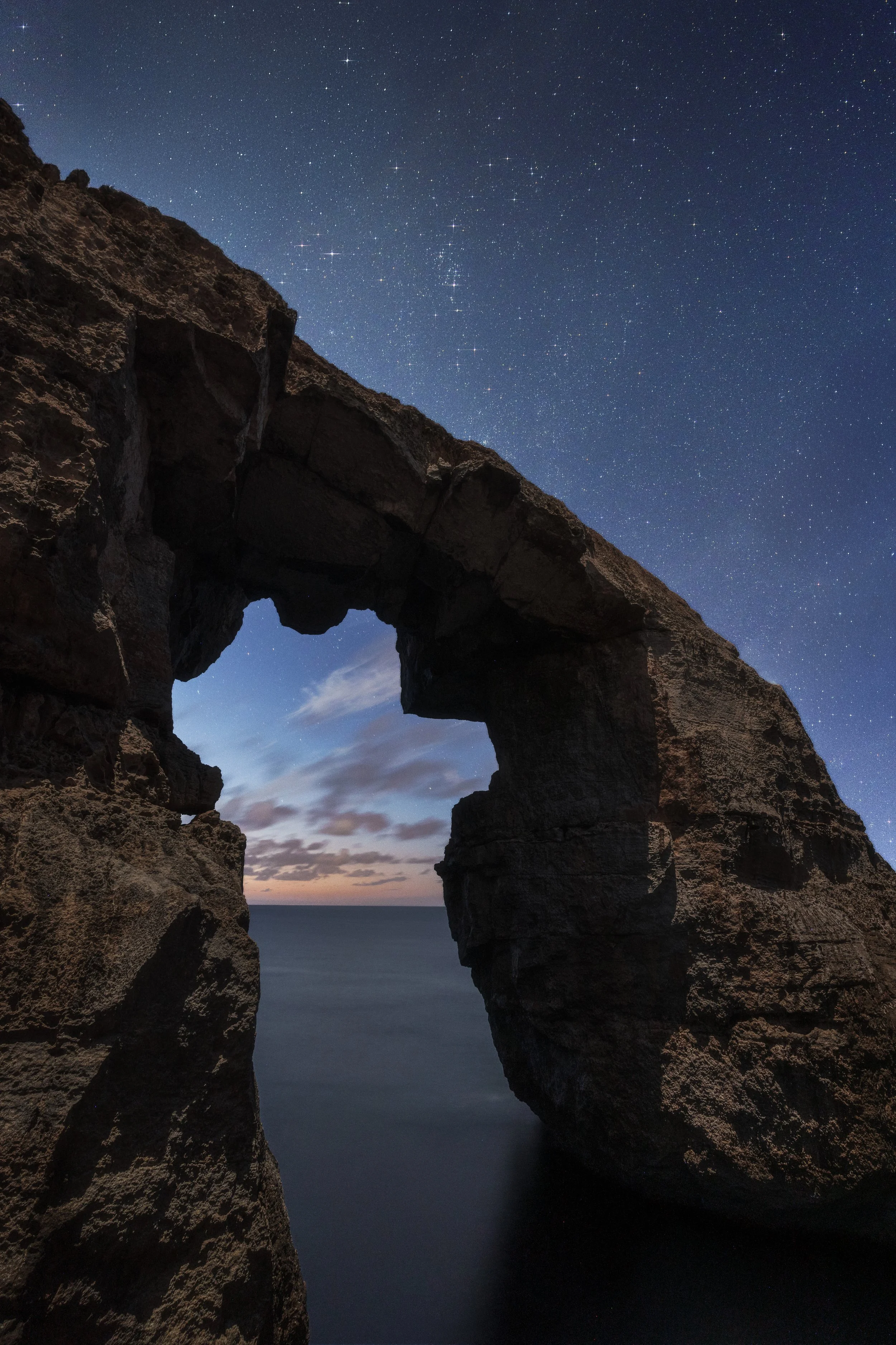 Nighttime view through a large rock arch over the ocean, with stars visible in the sky and some clouds near the horizon.
