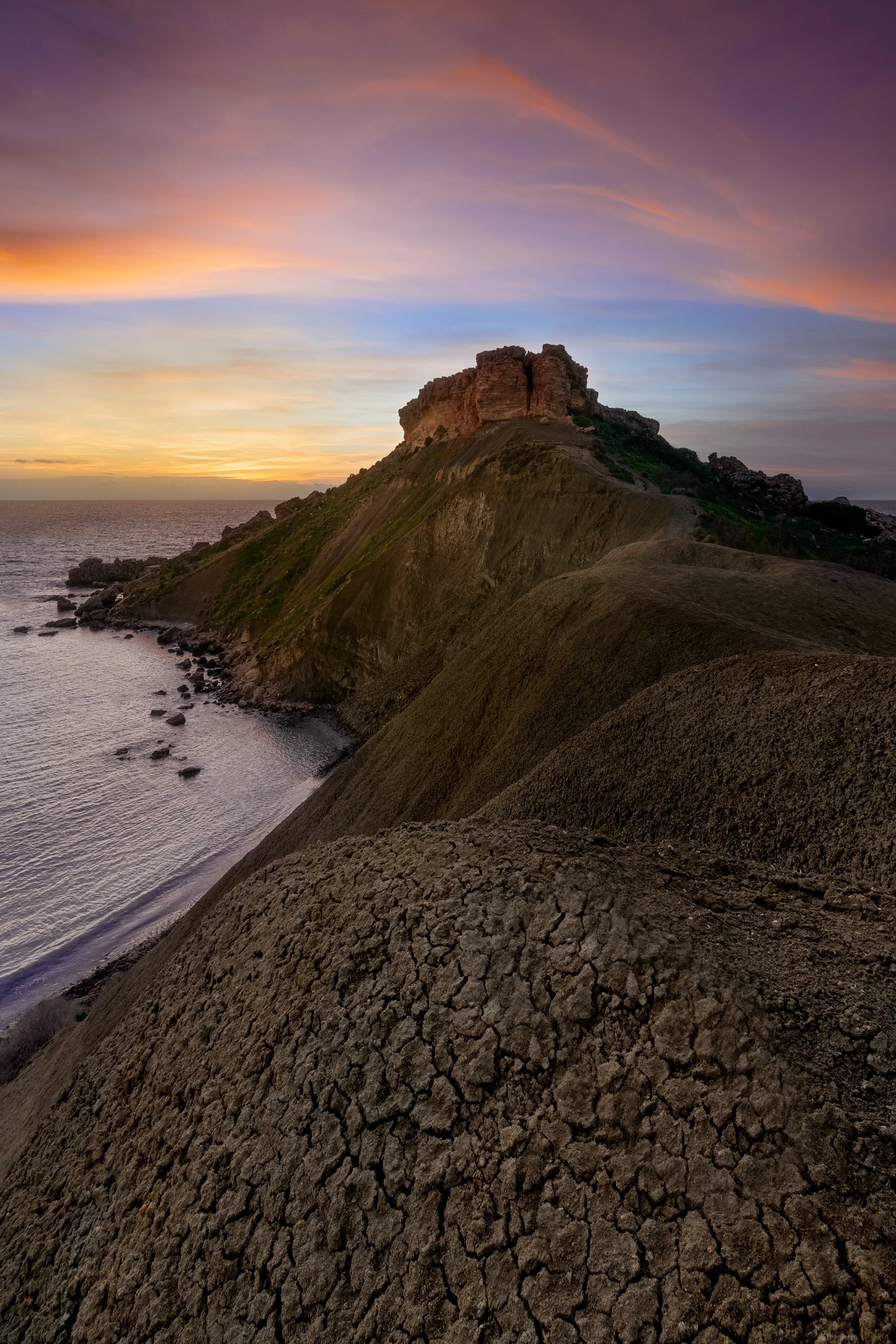 Sunset over a rugged cliffside with dry, cracked soil in the foreground, leading up to a rocky formation at the top of the cliff with an ocean background.