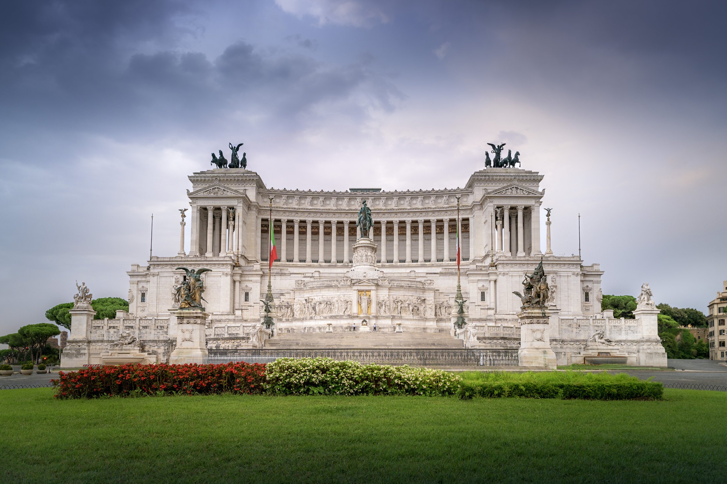 A large, ornate monument with white marble, featuring multiple statues and sculptures, including chariots on top, situated in a landscaped area with grass and flowers, under a cloudy sky.