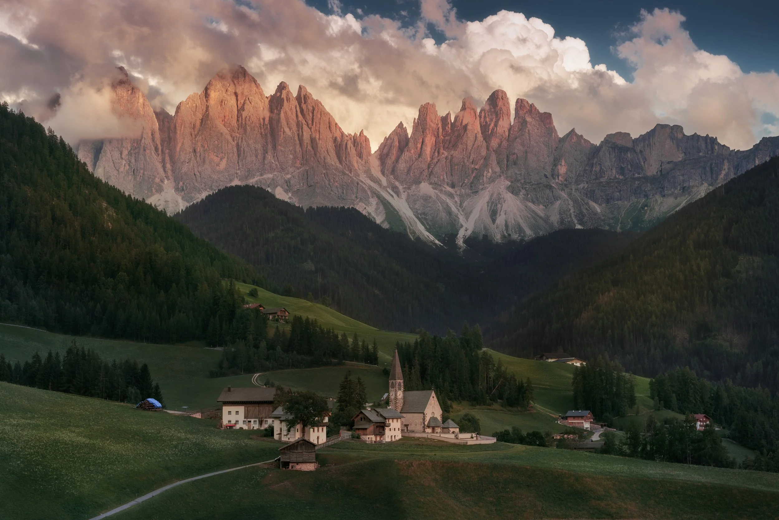 Scenic view of a small village with houses and a church at the base of green hills and towering mountains with clouds overhead.