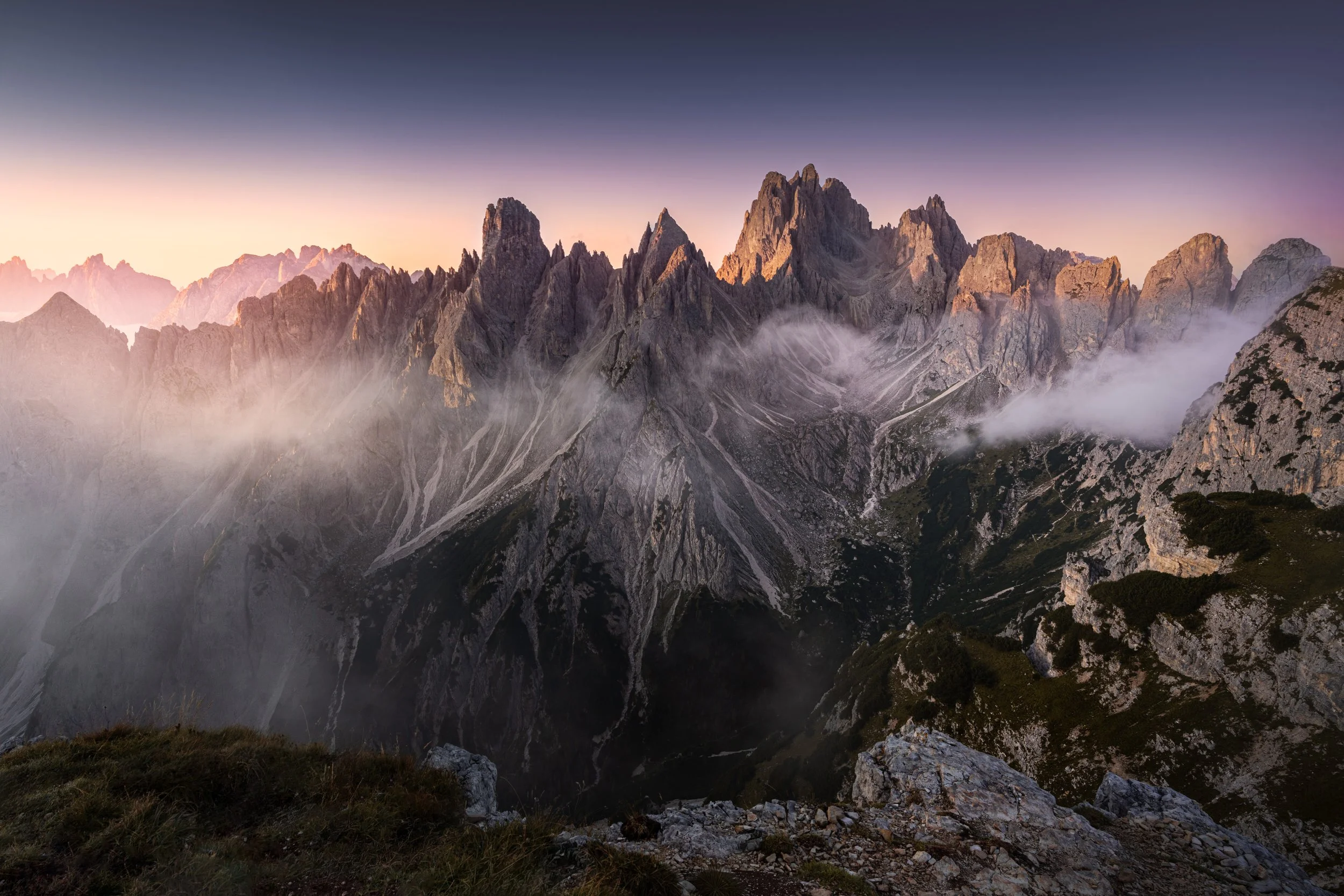 Sunrise over jagged mountain peaks with mist and clouds in the valleys.