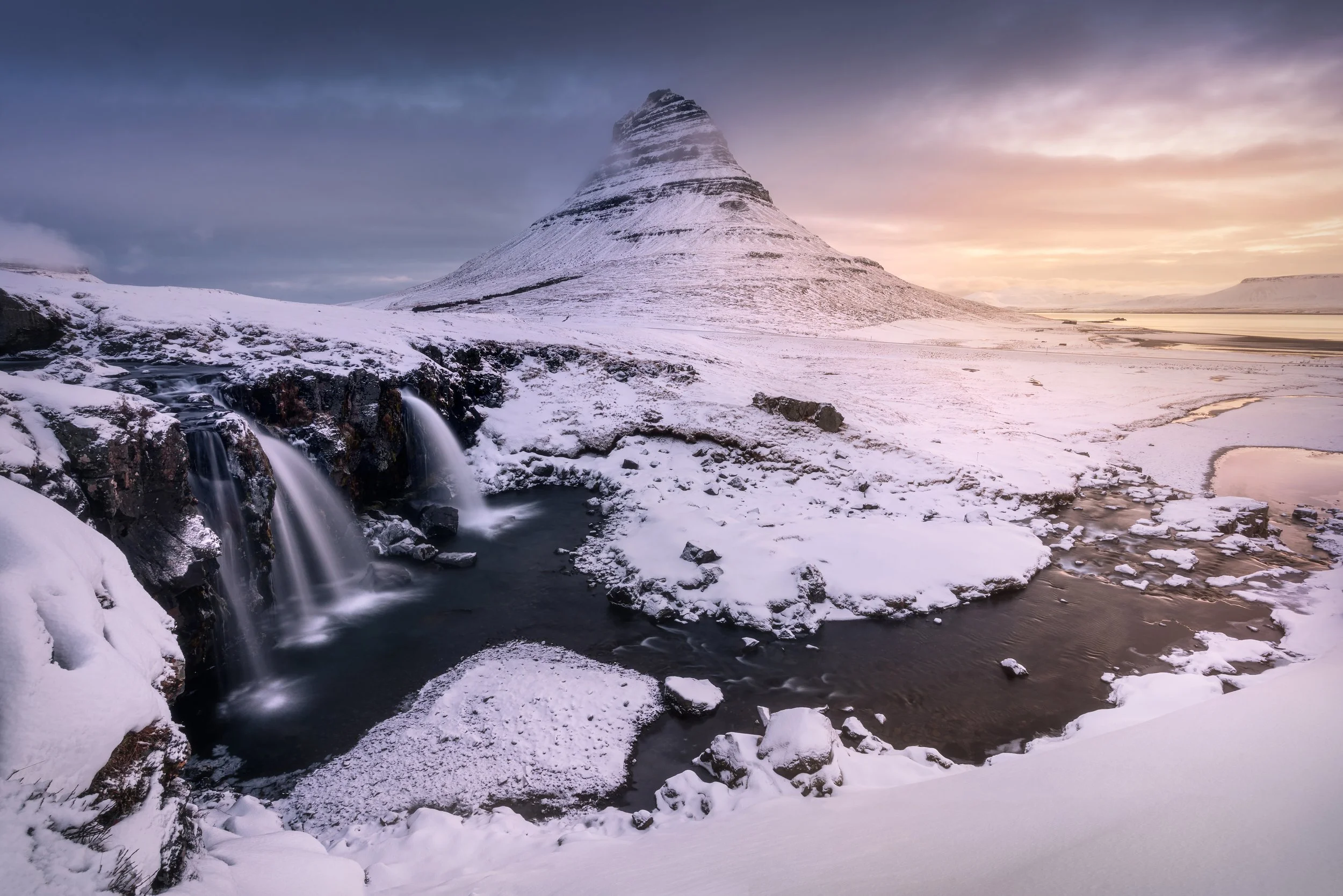 Snow-covered landscape with a waterfall in the foreground and a distinctive mountain in the background during sunset.