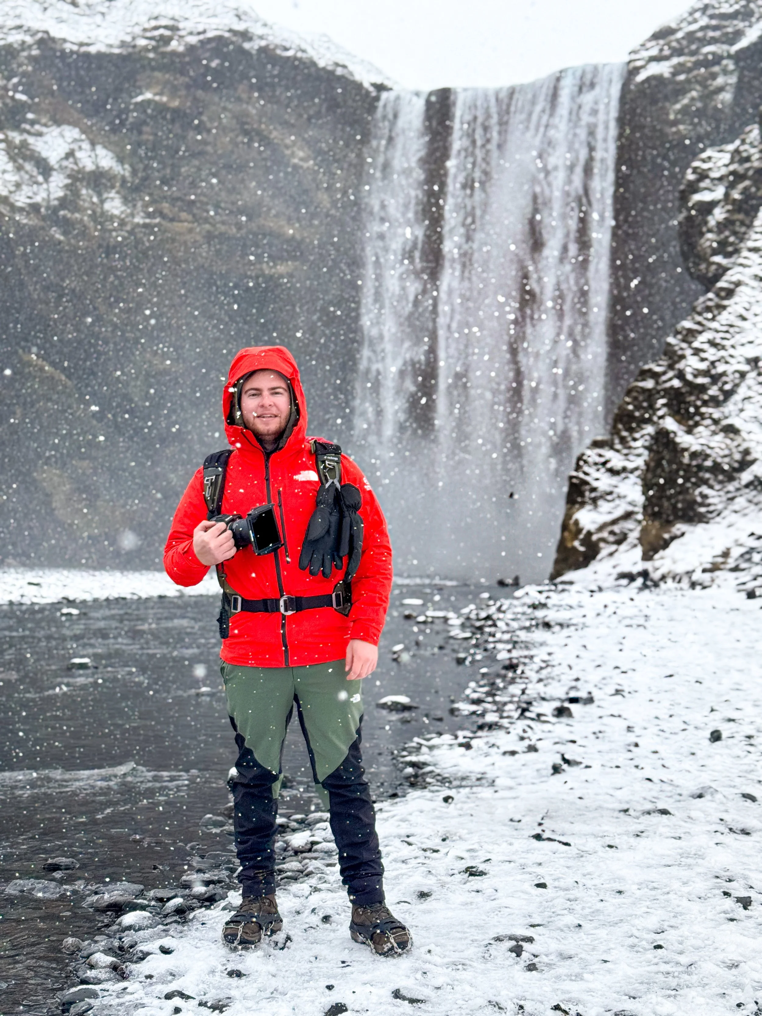 A man in a red waterproof jacket, green and black snow boots, and green and black pants stands in the snow near a waterfall. He is holding a camera, and snow is falling around him.