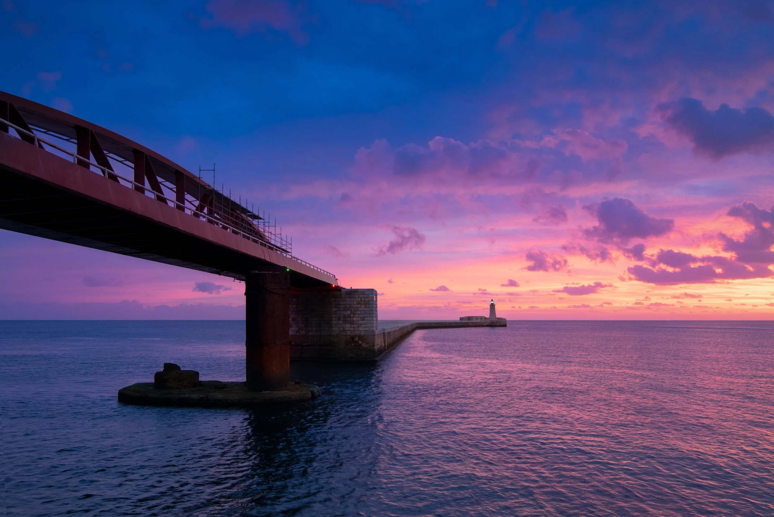 A pier extending into the ocean at sunset with a lighthouse at the end, colorful pink and purple sky with scattered clouds.