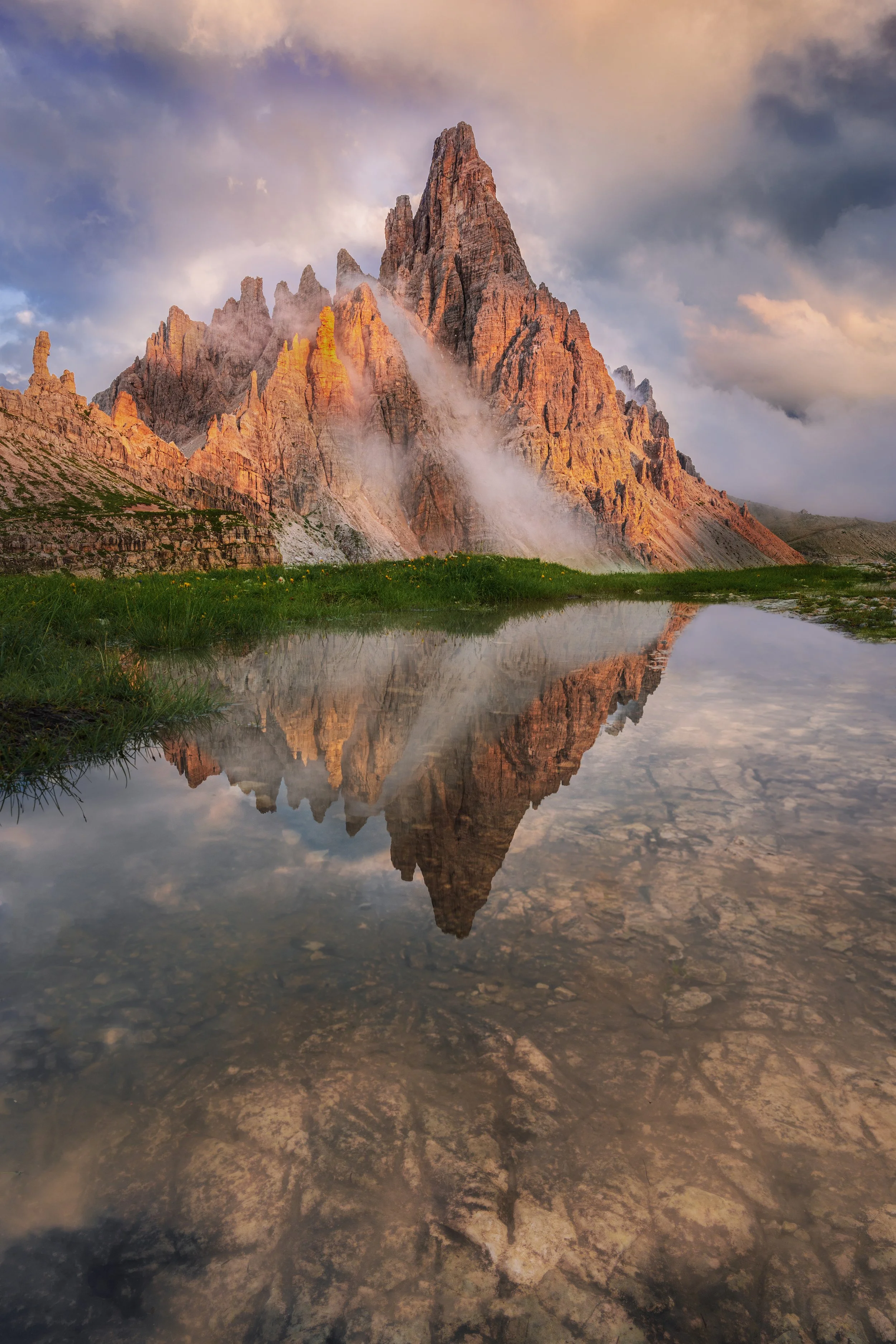 A mountain with sharp peaks reflected in a still body of water, with clouds in the sky.