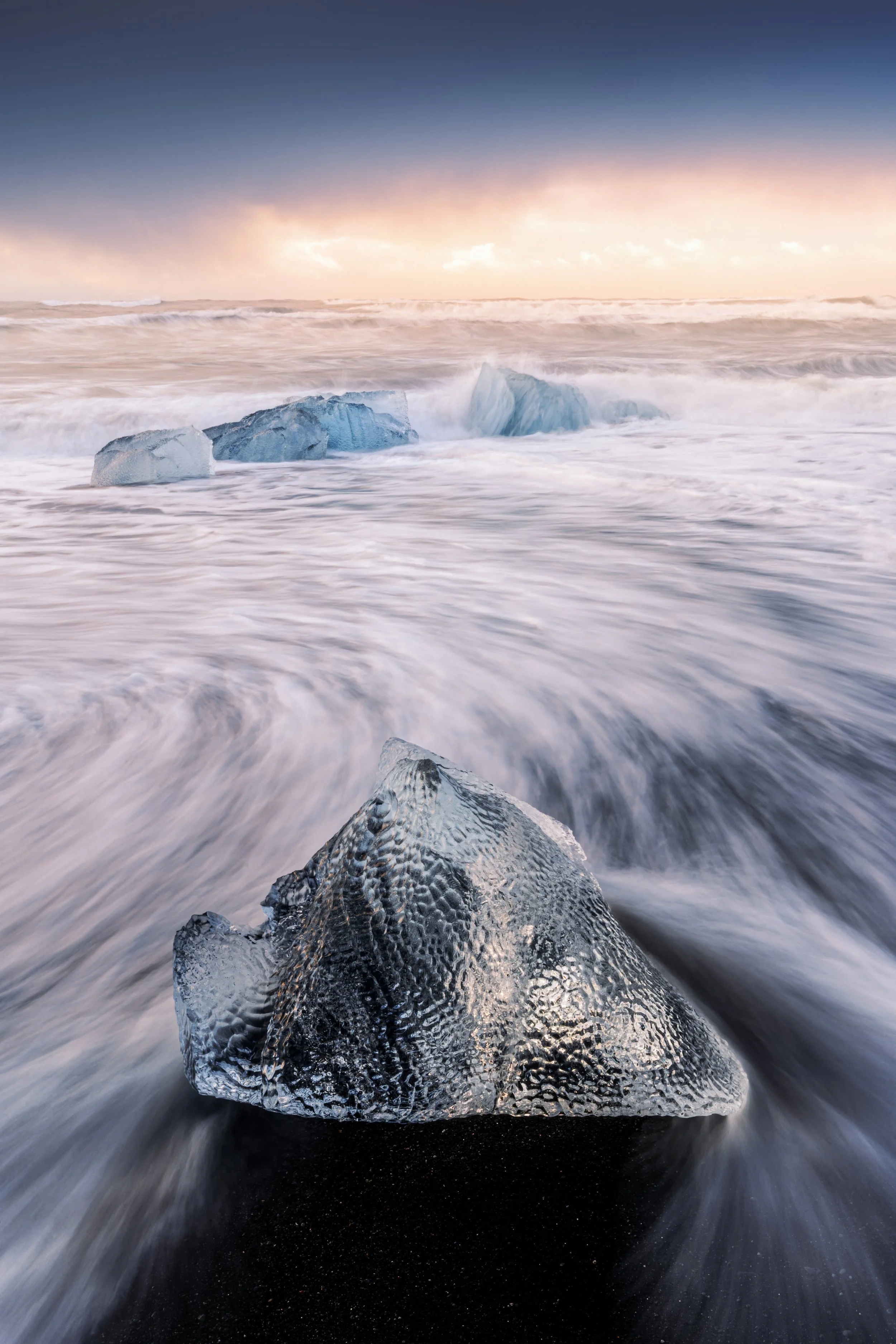 Iceberg on a black sand beach with waves crashing around it, under a colorful sky at sunset.
