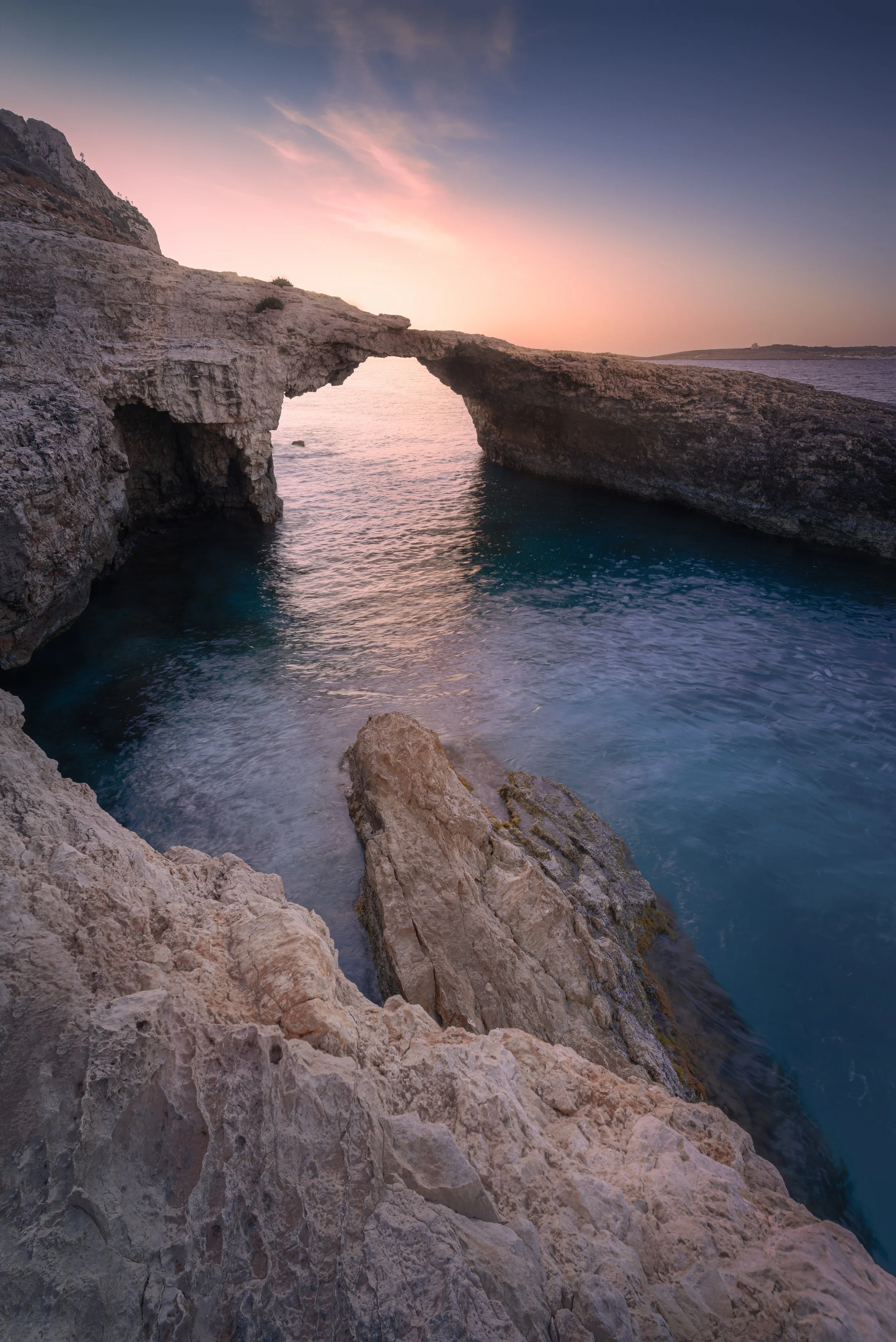 A natural rock arch over the ocean at sunset with pink and blue hues in the sky.