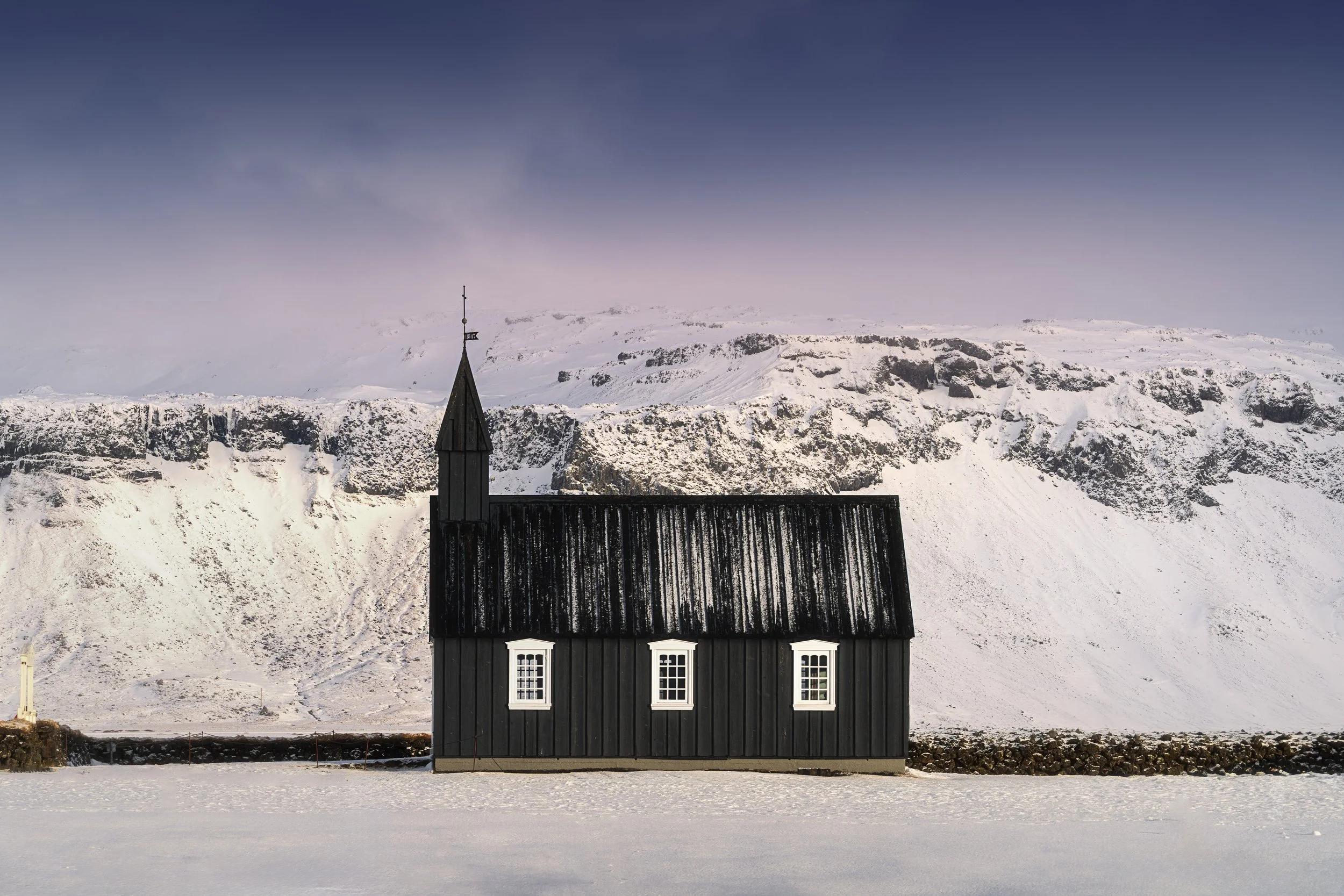 A black wooden church with a steeple is situated in a snow-covered landscape with mountains in the background.