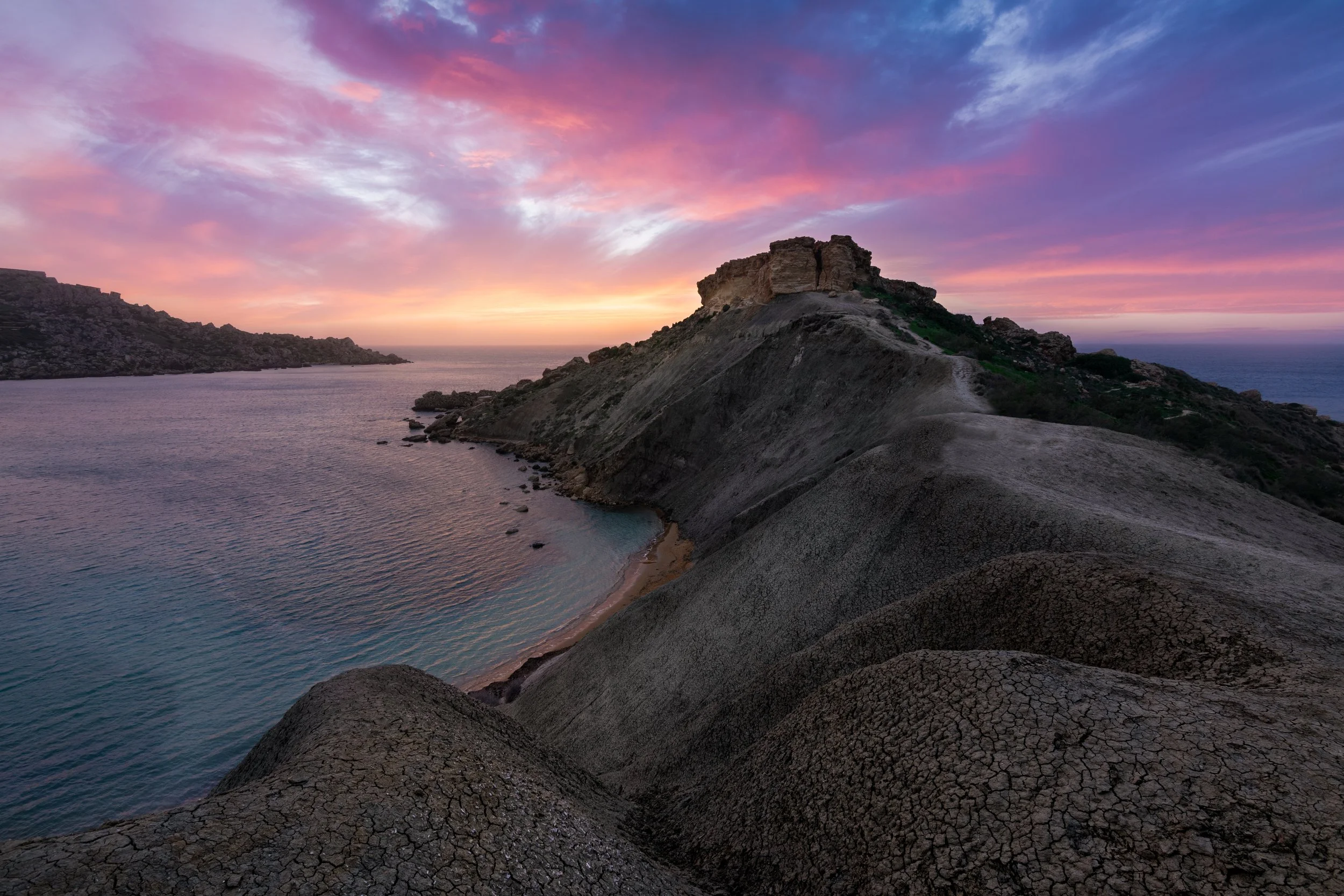 A coastal landscape at sunset with a rocky hill in the foreground, a small beach at the base, and a colorful sky with pink, purple, and orange hues.