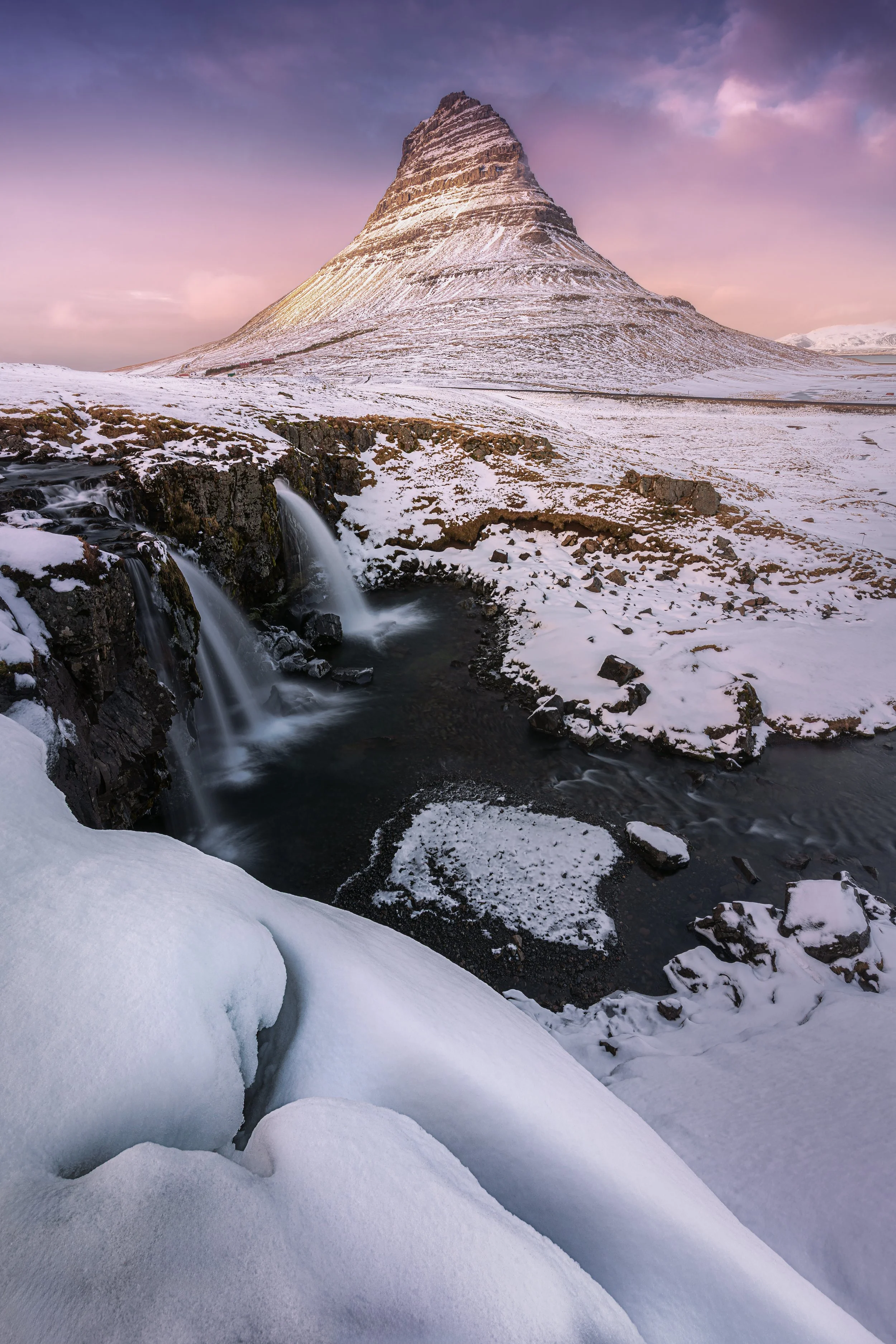 Snow-covered mountain with a pointed peak, a small waterfall flowing into a river, snow-covered rocks, and a colorful sky at sunset.