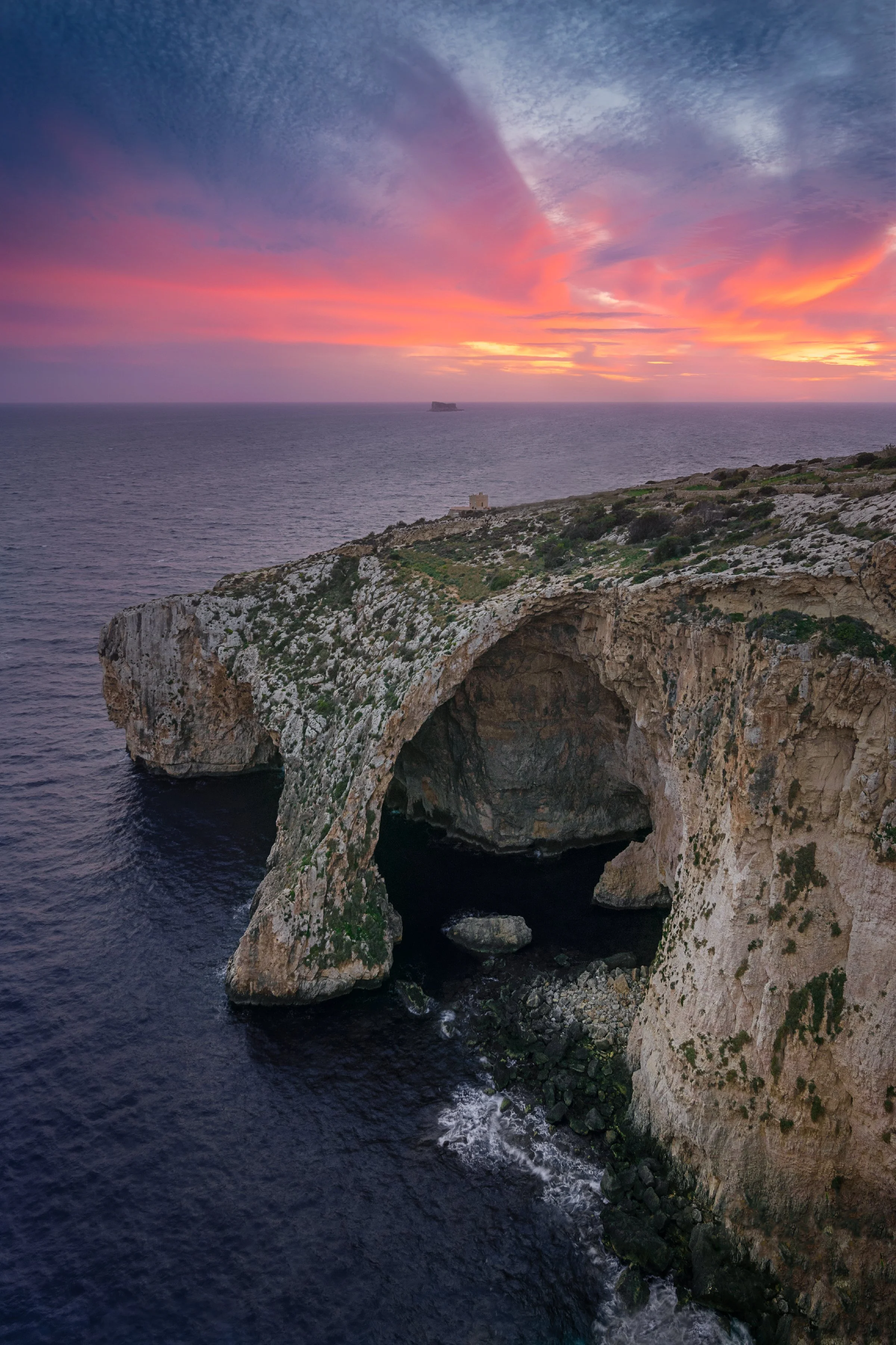 Aerial view of a coastal cliff with a natural arch, overlooking the ocean at sunset with pink and orange clouds in the sky.