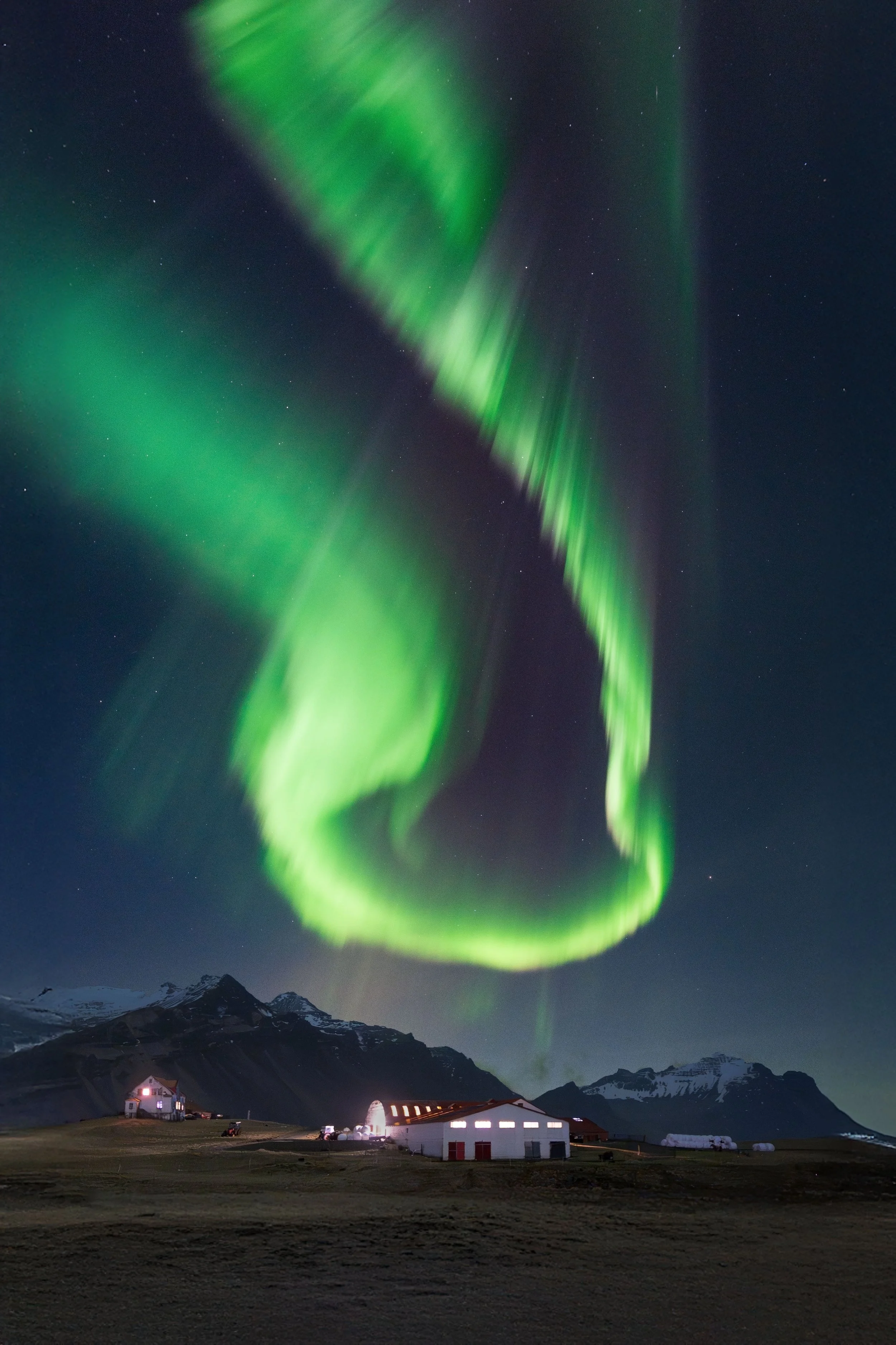 Northern lights illuminating the night sky over a rural farm with barns and mountains in the background.