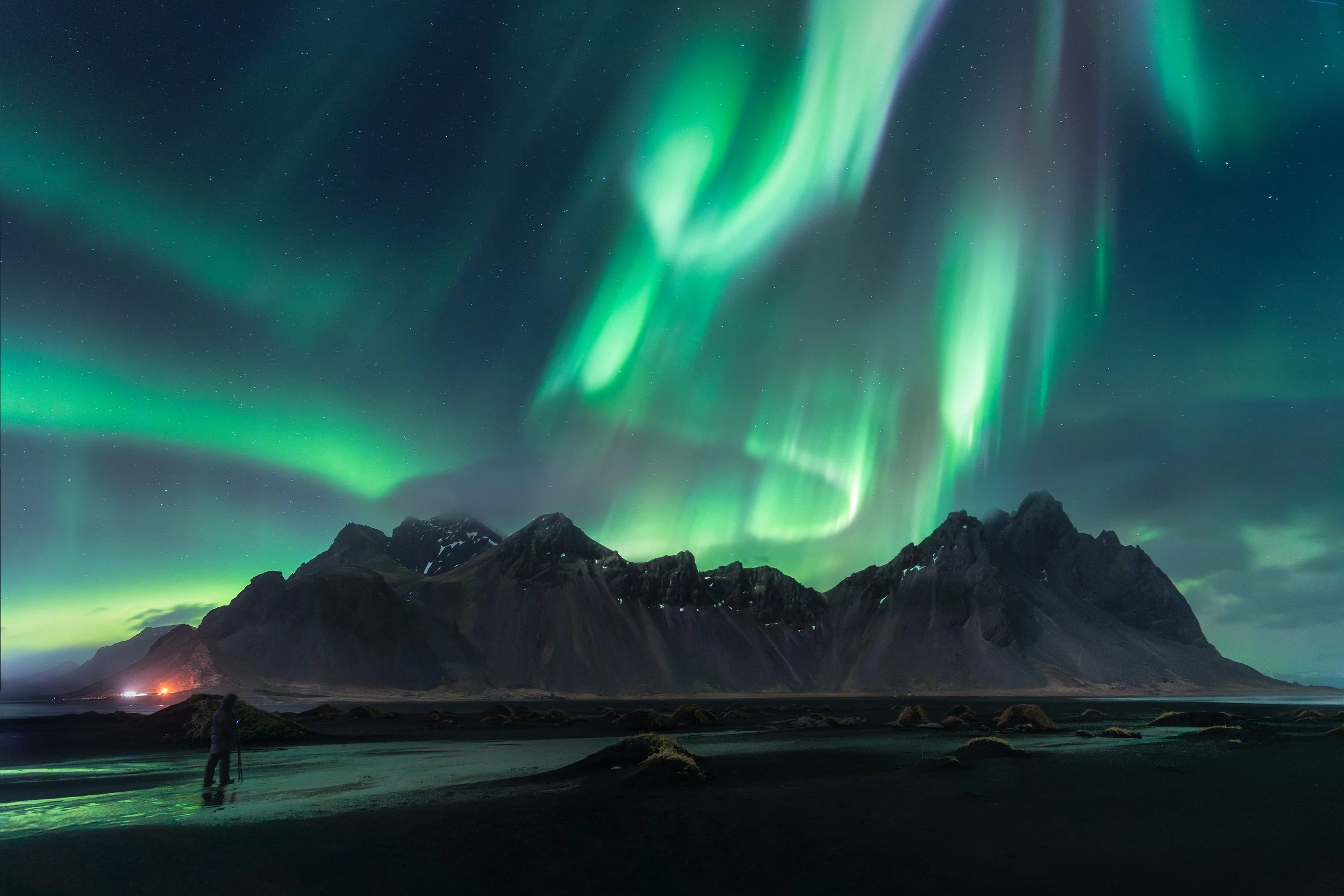 Northern lights illuminating the night sky over a mountain landscape with a person standing near water in the foreground.