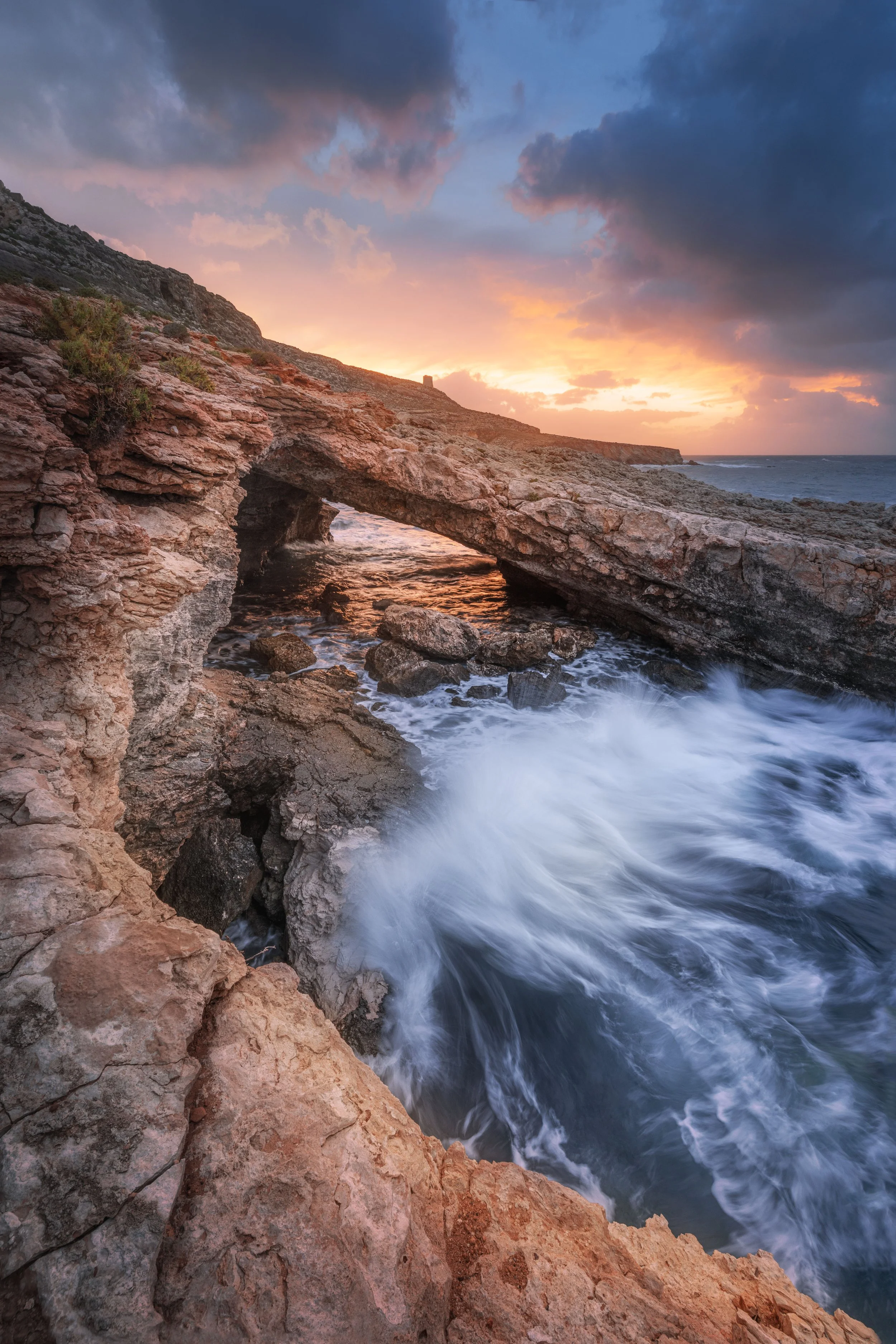 Coastal scene at sunset with waves crashing against rocky cliffs and a partly cloudy sky.
