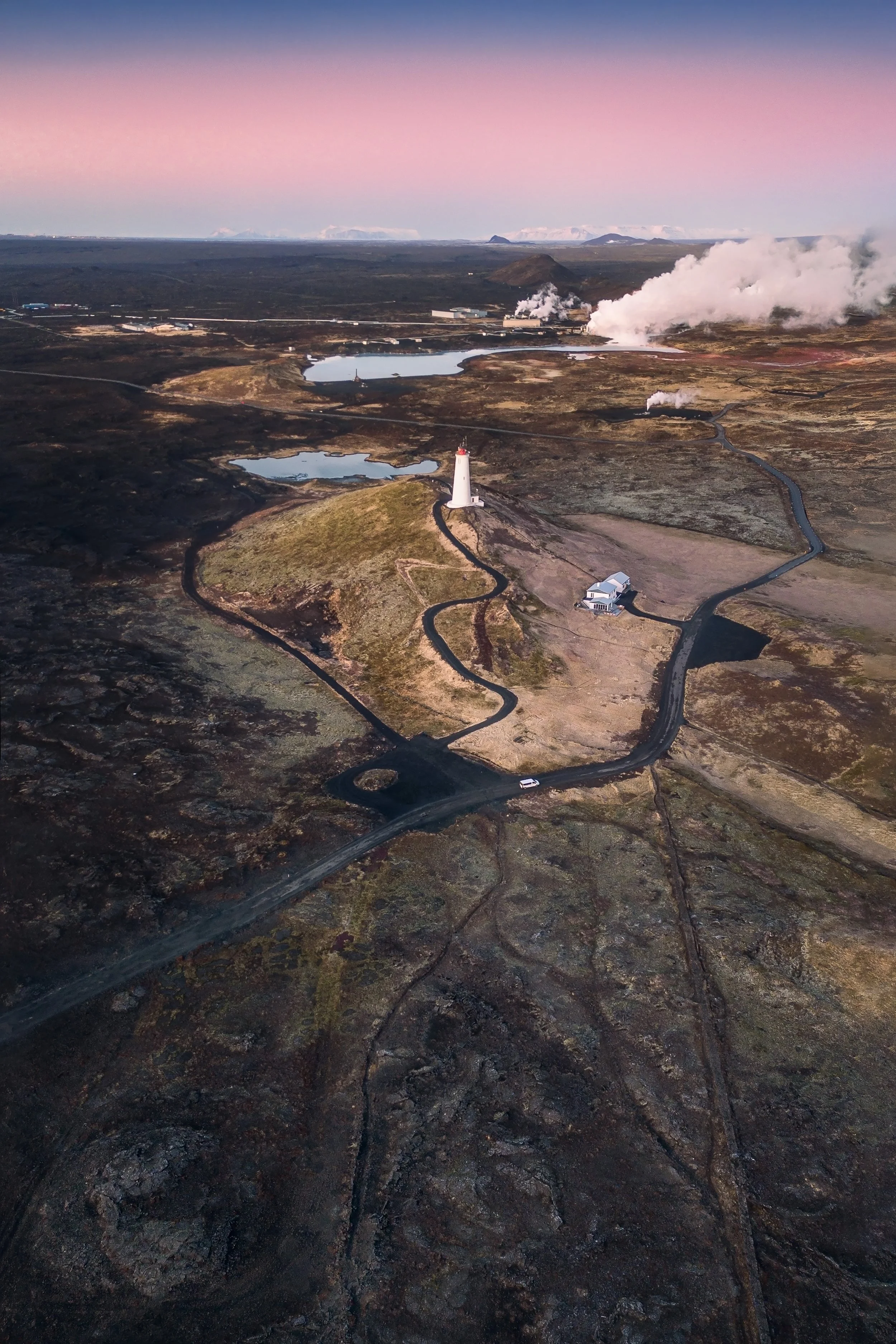 Aerial view of a volcanic landscape with a winding road, lighthouse, and small building, steaming vents, and volcanic craters at sunrise or sunset.