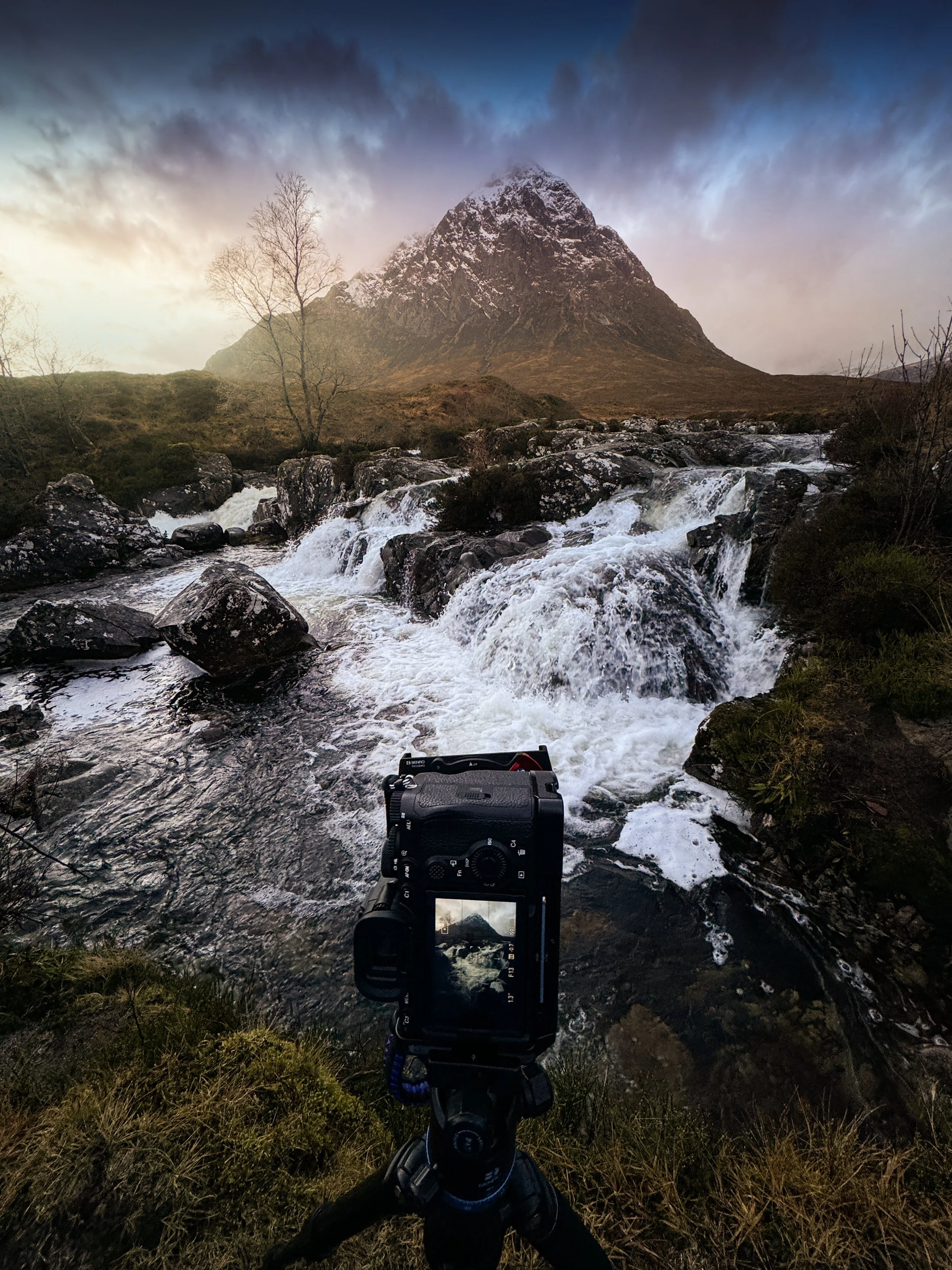 Camera on tripod capturing a mountain scene with a rushing stream in the foreground, leafless trees, and snow-capped peaks under cloudy sky.
