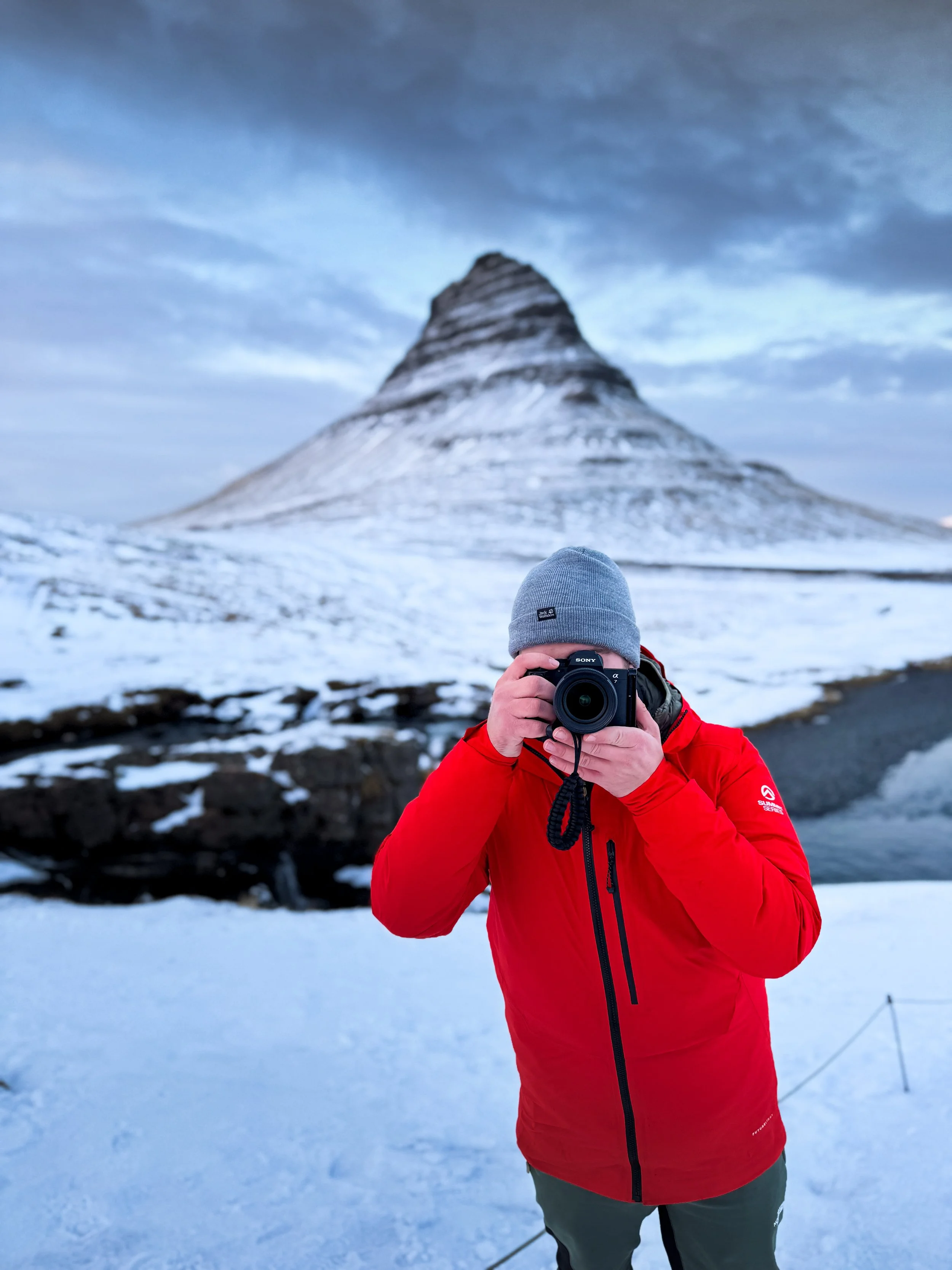 Person in red jacket and gray beanie taking a photo with a camera in a snowy landscape with a mountain in the background.