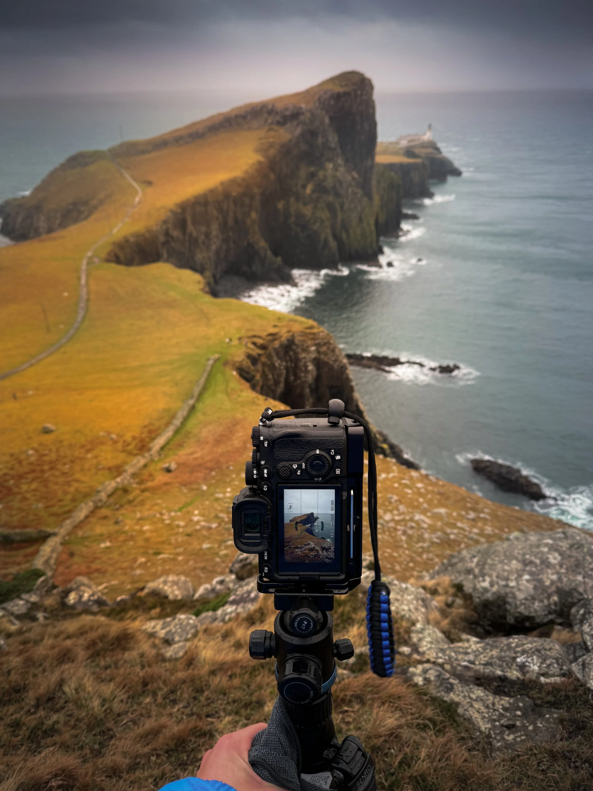 A camera on a tripod capturing a view of rugged coastal cliffs and the ocean, with a lighthouse on a distant point, overcast sky.