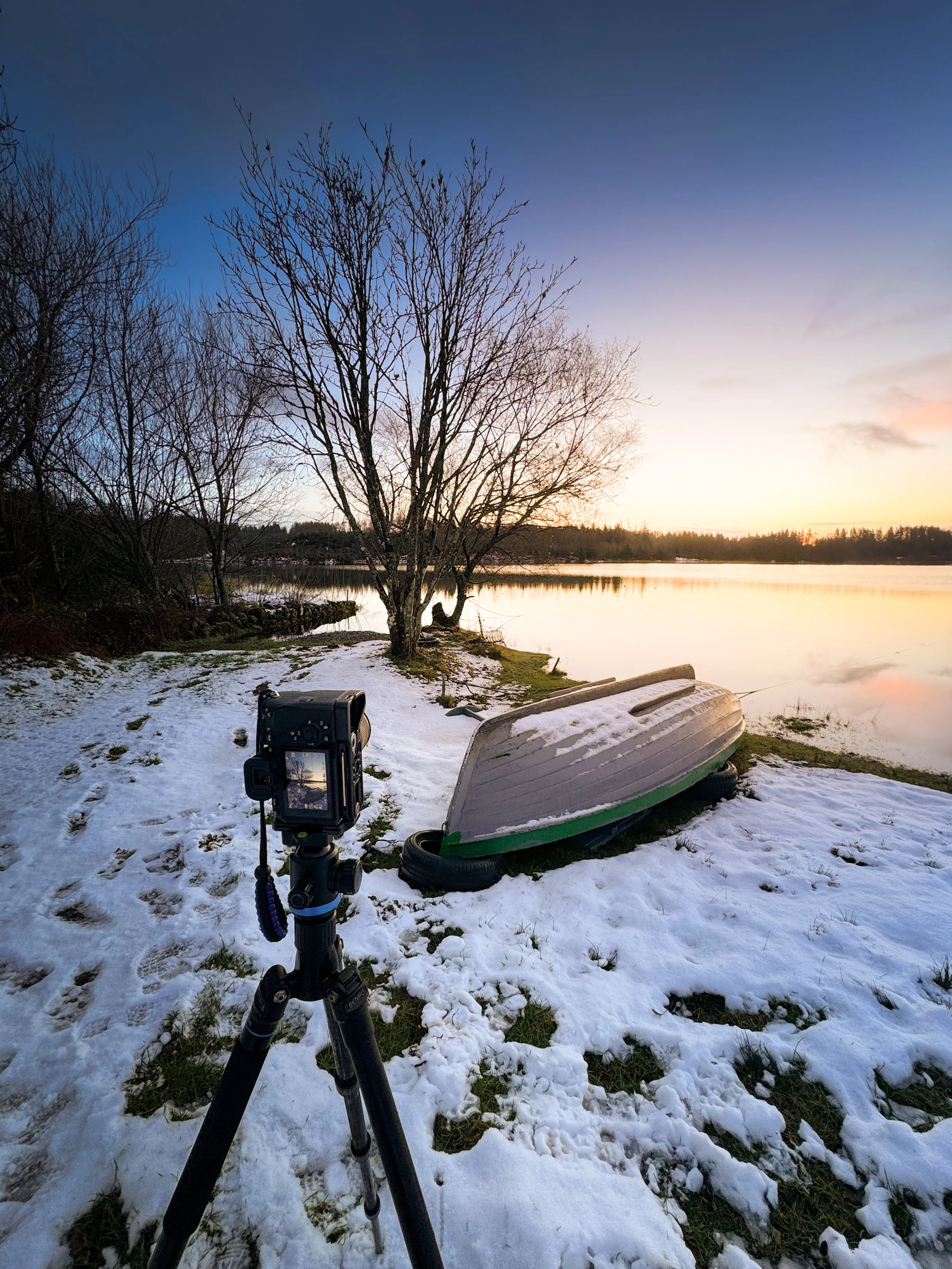 A landscape scene at sunset or sunrise by a lake with snow-covered ground, a leafless tree, a boat turned upside down on the shore, and a camera on a tripod capturing the scene.