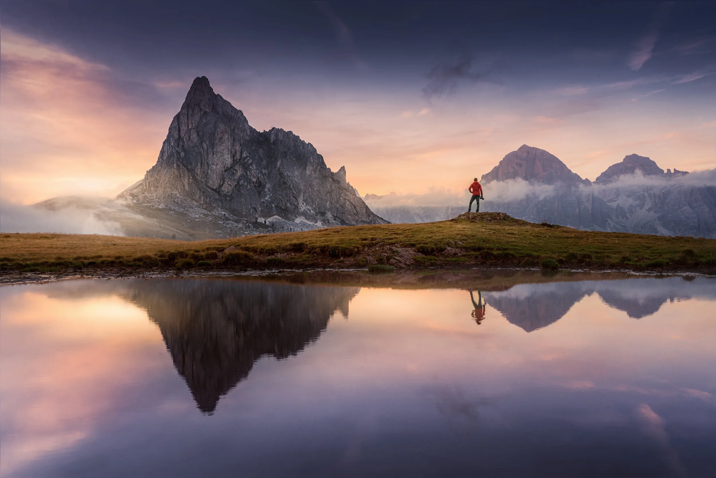A person standing on a grassy hilltop overlooking a reflective body of water with mountains in the background at sunset.