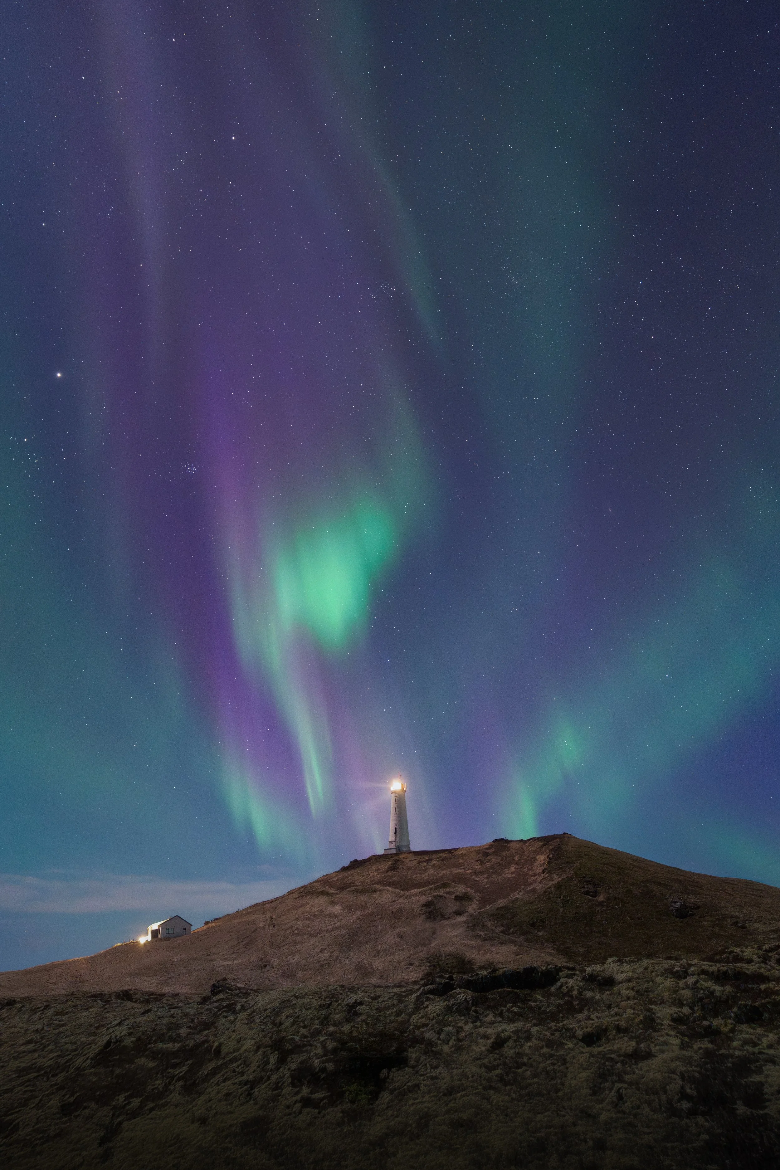 Night sky illuminated by aurora borealis with a lighthouse on a hill.
