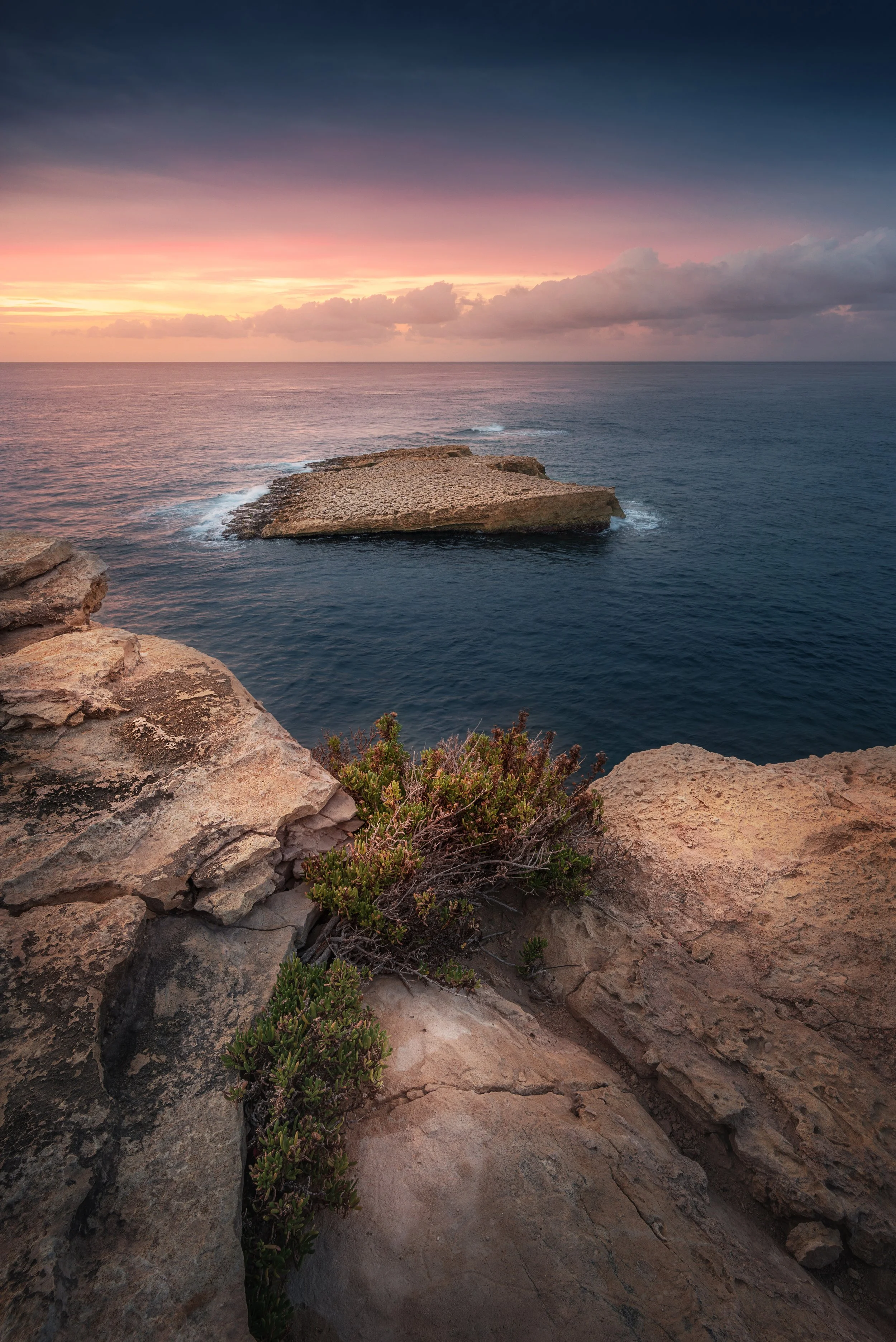 A scenic view of a rocky coastline at sunset, with a large rock formation in the ocean and a partly cloudy sky with pink and purple hues.
