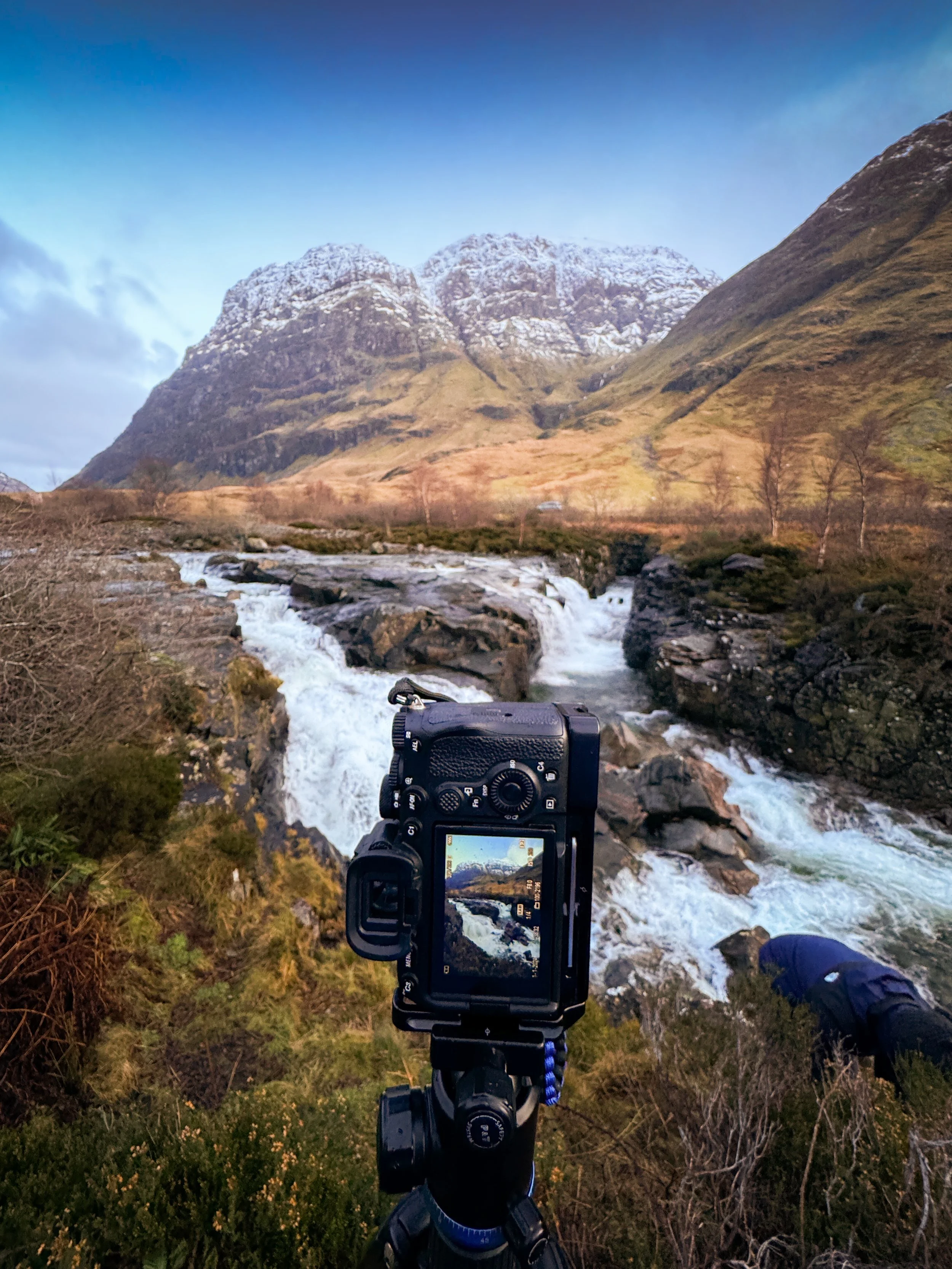 A camera on a tripod taking a photo of a waterfall in a mountainous landscape, with snow-capped peaks in the background.