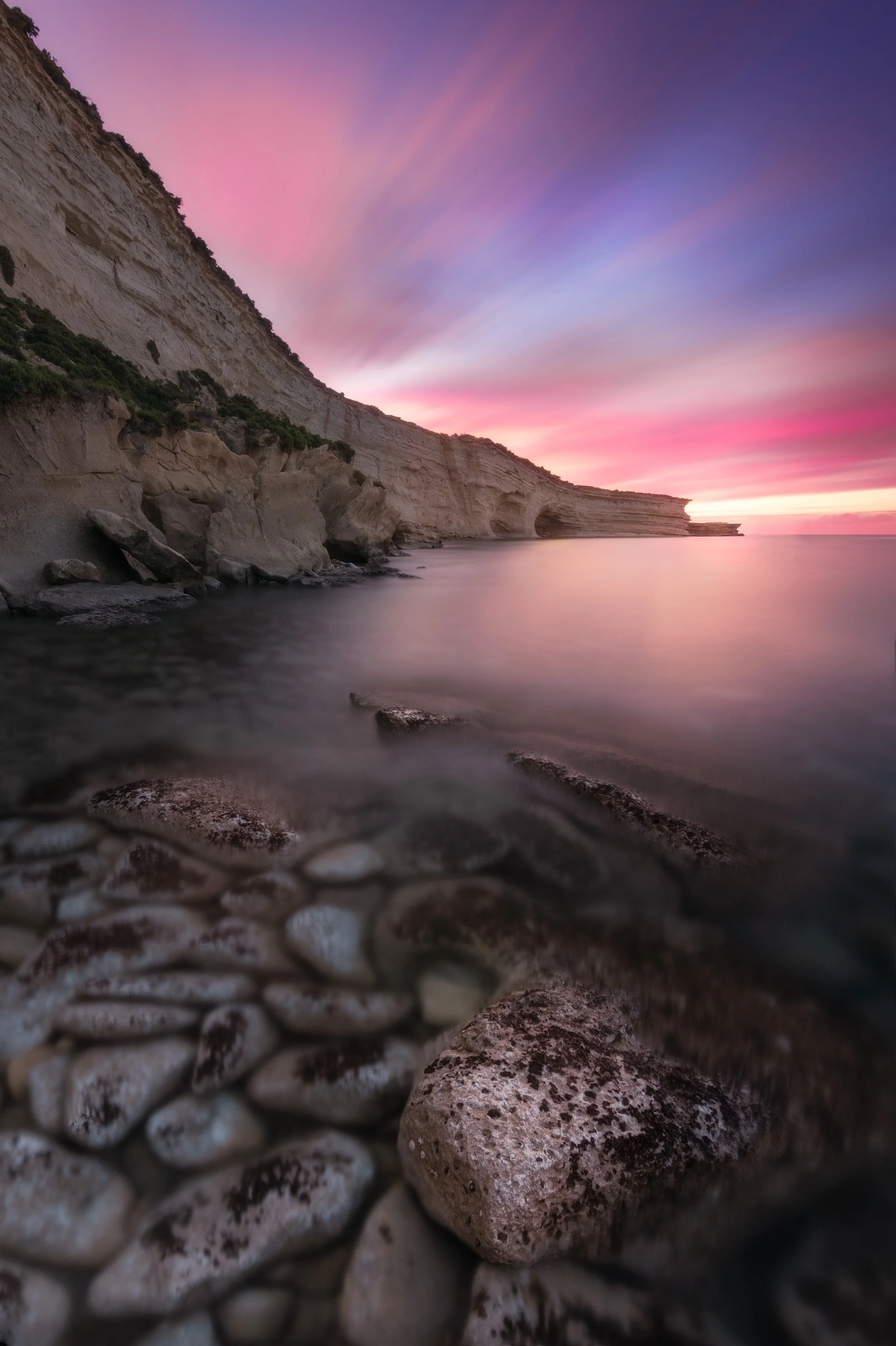 Sunset over rocky coastline with cliffs, calm water, and pink, purple, and blue sky.