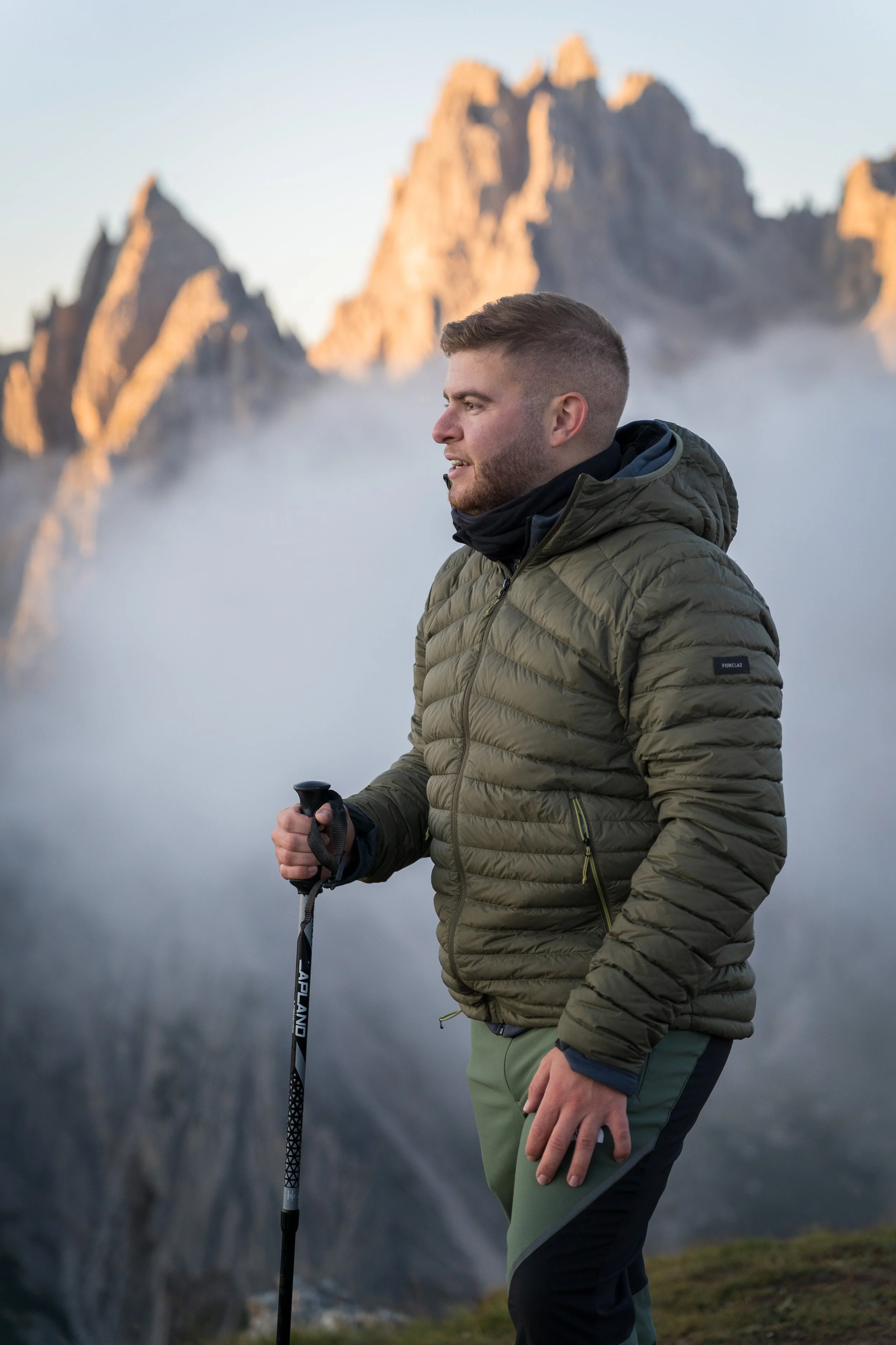A man standing outdoors in a mountainous area with fog, holding a walking stick, wearing a green insulated jacket and dark pants.