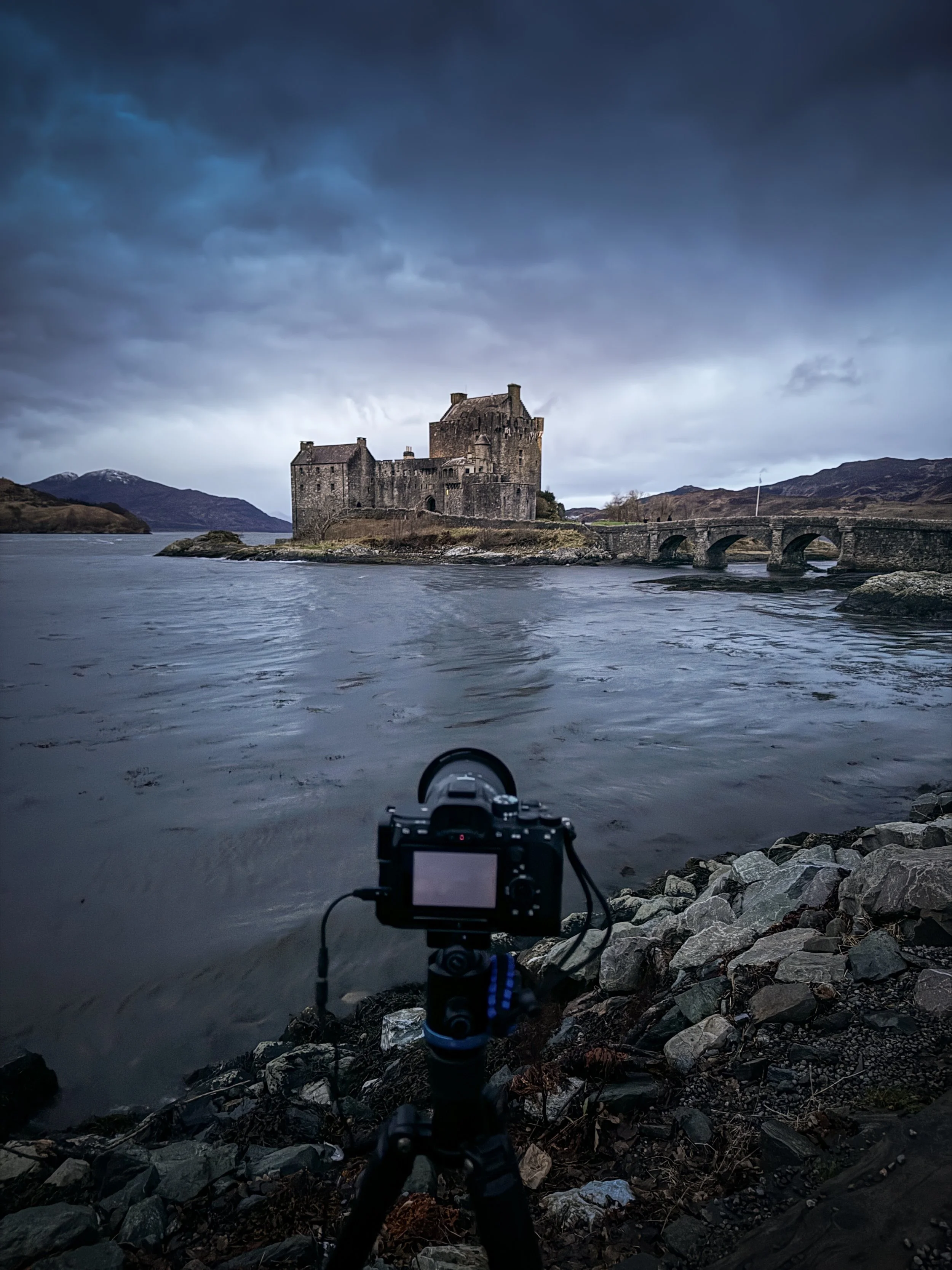 A camera on a tripod positioned on rocky shoreline takes a photo of a medieval castle on a small island, connected by a stone bridge, under a cloudy sky.