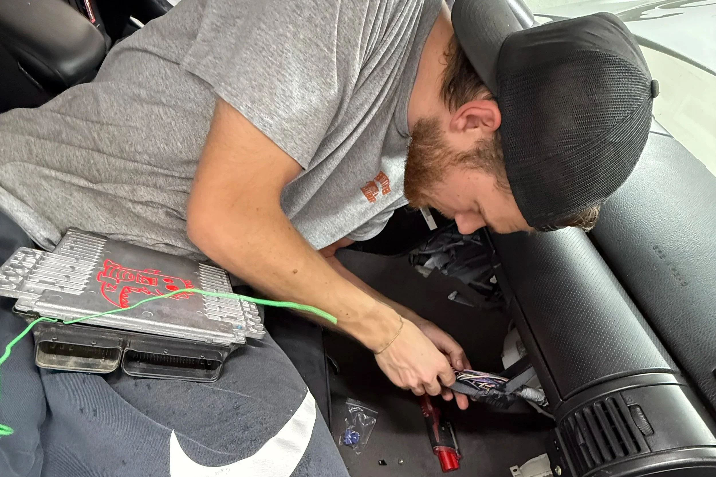A man wearing a gray t-shirt and black cap working on a car's dashboard, with electronic tools and wires visible nearby.