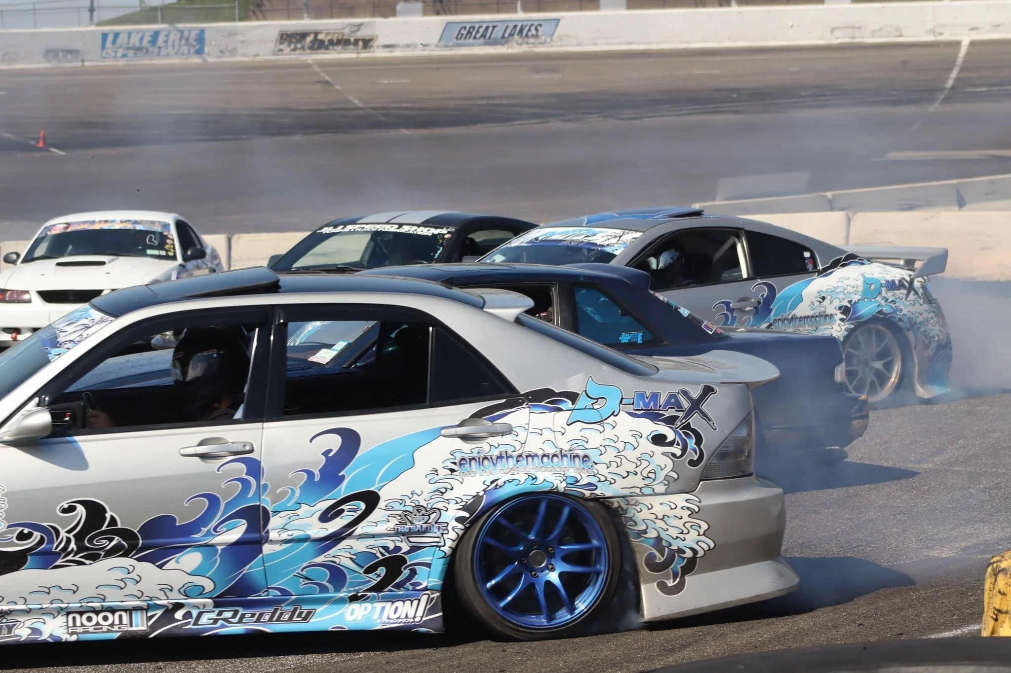 Multiple race cars on a track, with a silver car featuring a blue cloud and wave design in the foreground. Smoke is coming from the rear tires of some cars, indicating drifting or racing maneuvers.