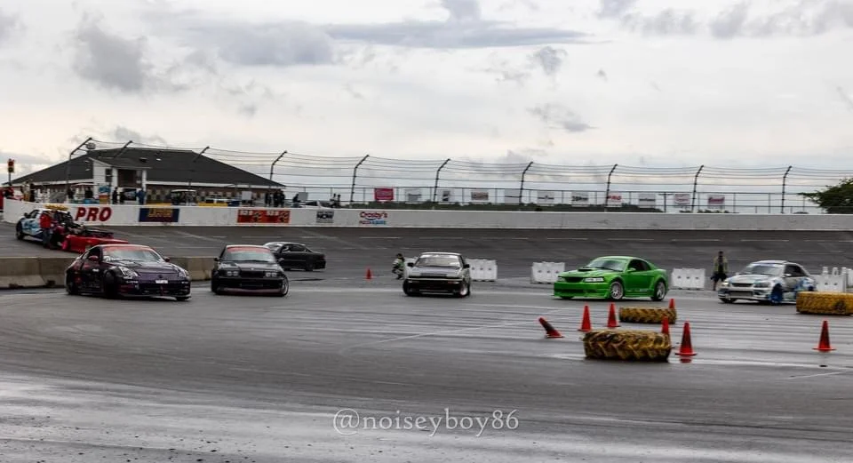 Several sports cars parked on a racetrack behind orange cones and tire barriers under a cloudy sky, with a racetrack grandstand in the background.
