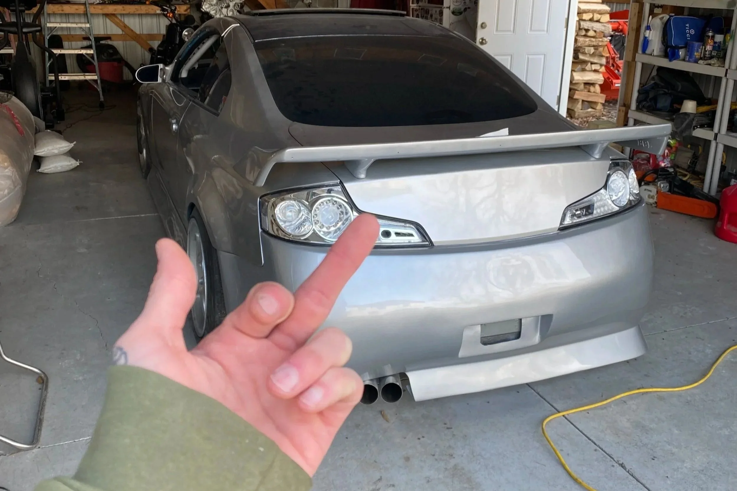 A silver sports car with a large rear spoiler is parked inside a garage. A person is in front of the car, making a gesture with middle finger and index finger extended. The garage contains shelves with tools and supplies, as well as cardboard boxes and a red fuel container.