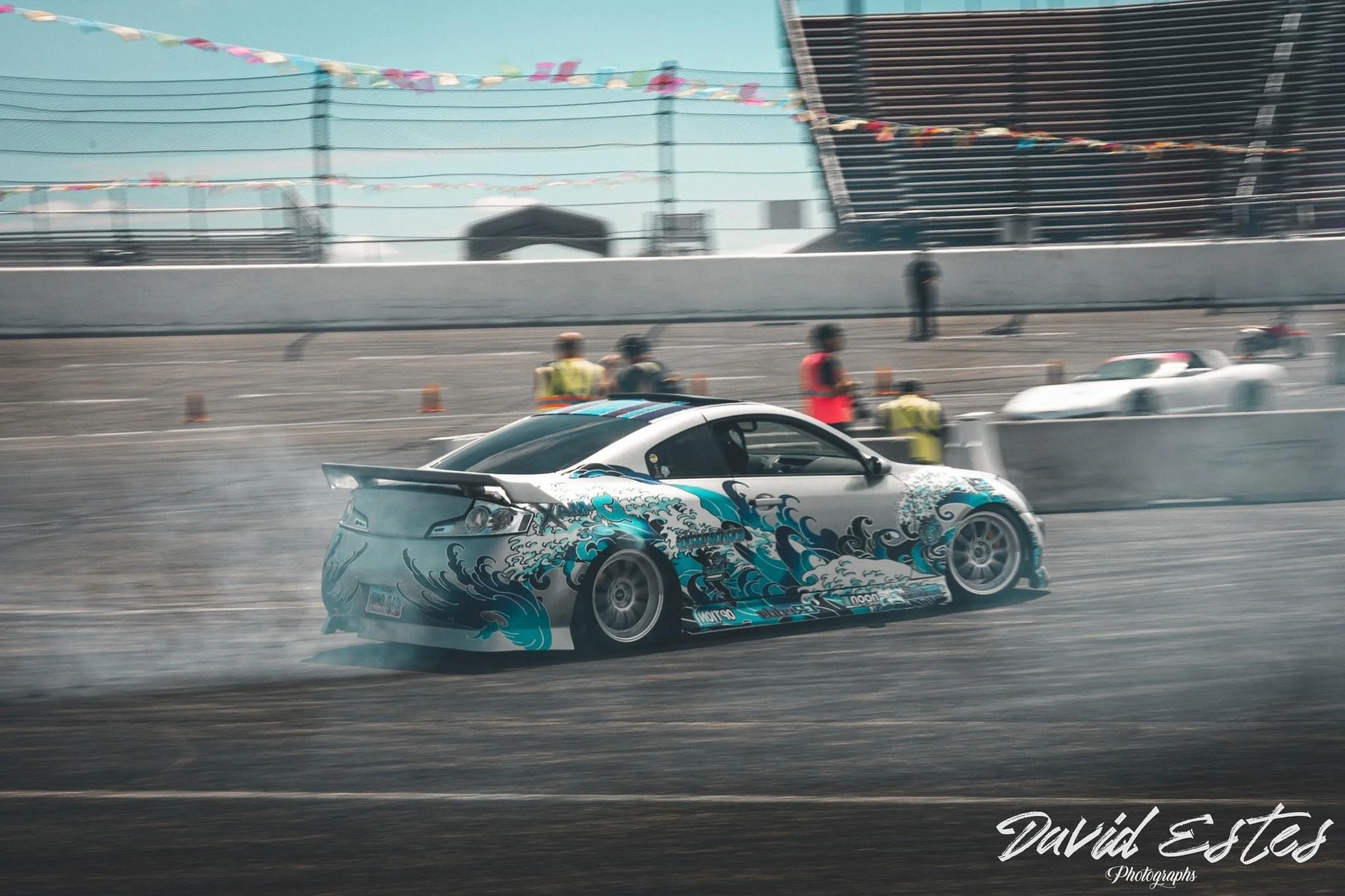 A race car with wave-themed graphics drifting on a race track during a motorsport event, with spectators and crew in the background.