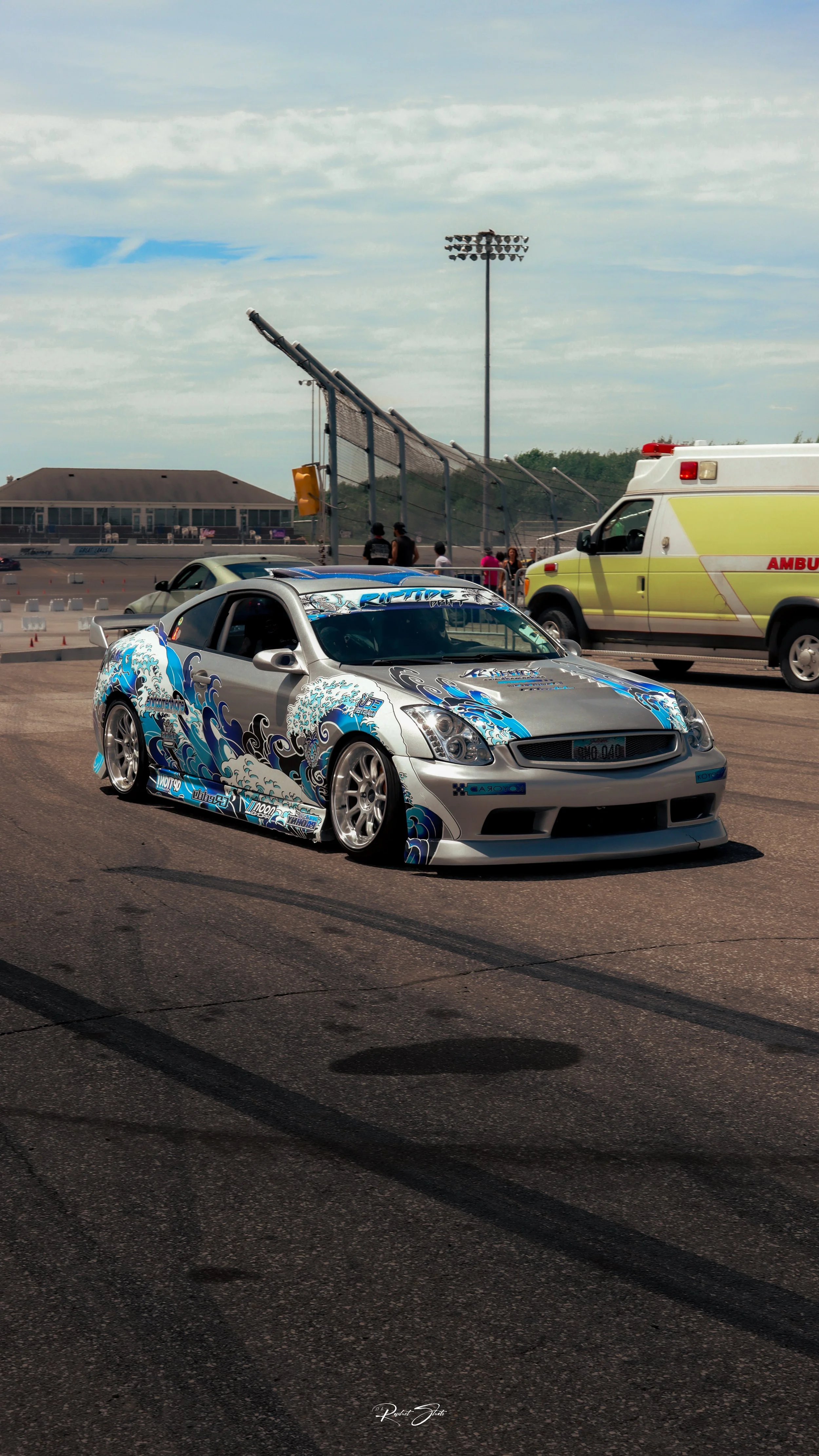 A silver sports car with blue and black wave-themed graphics drives on a racetrack, surrounded by an ambulance and a few spectators, with a light blue sky and clouds in the background.