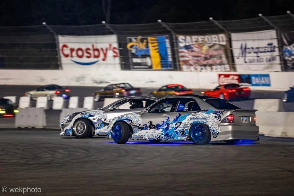 Two modified race cars, one silver with blue decorative waves and the other silver, side by side on a race track at night, with other cars and advertising banners in the background.