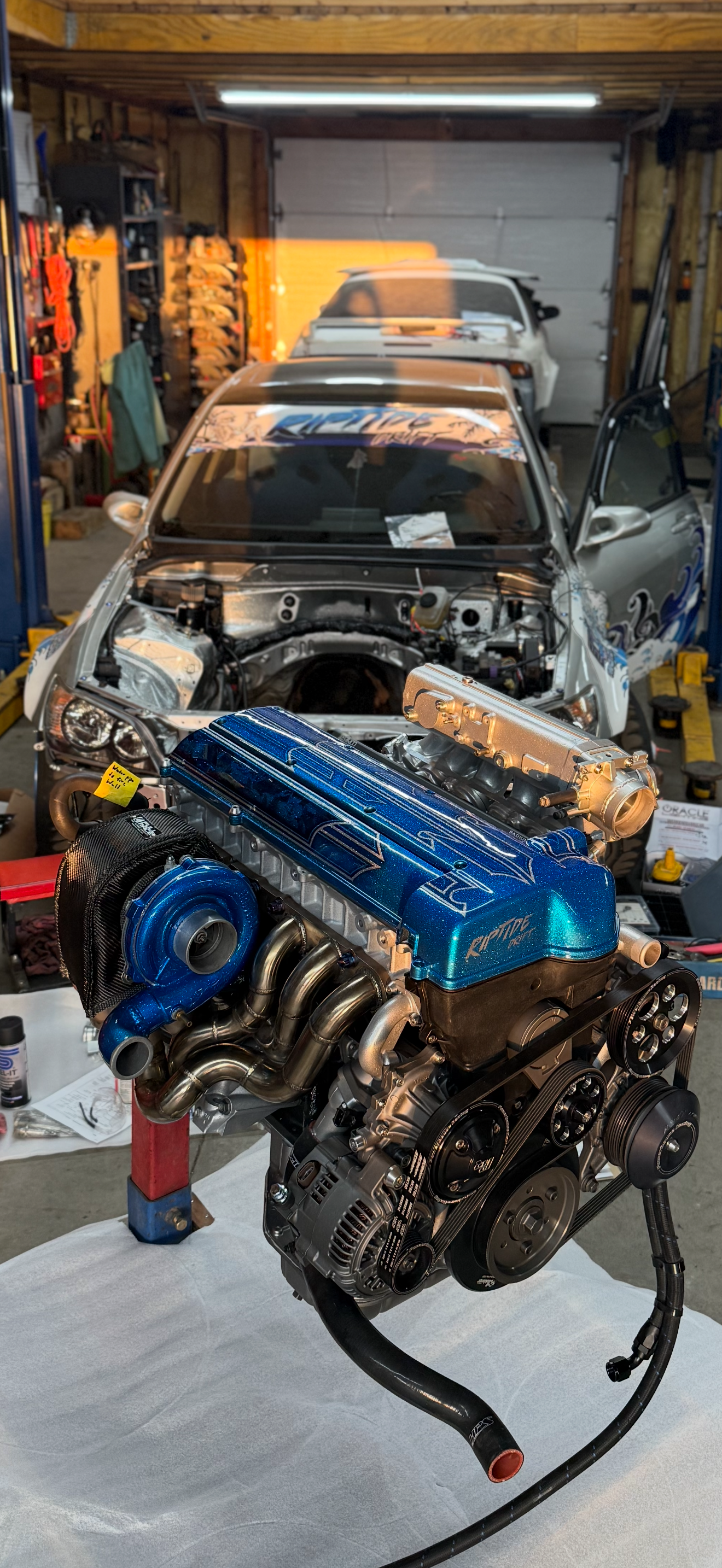 A detailed view of a high-performance engine in a garage with two cars, one in the background and one with a partially disassembled front, suggesting a car repair or modification project.