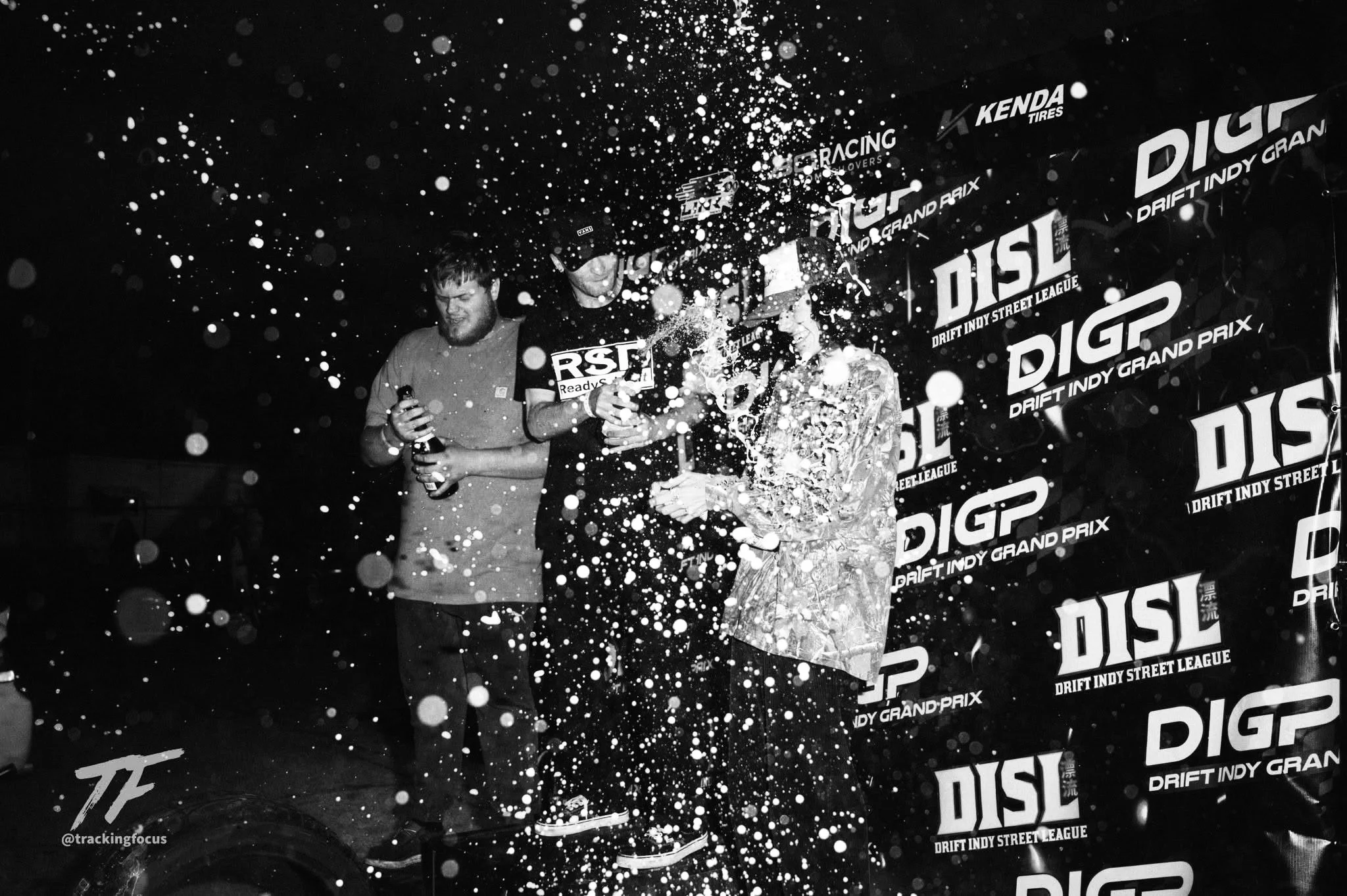 Three men celebrating on a podium with confetti in the air, at the Drift Indy Street League event, with a backdrop displaying the event logo.