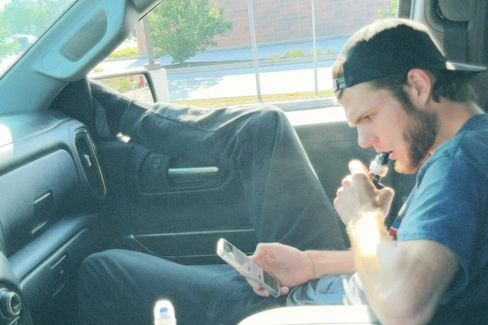 A young man with a beard, wearing a blue shirt and a backwards baseball cap, sitting in the passenger seat of a vehicle, using a smartphone and vaping.