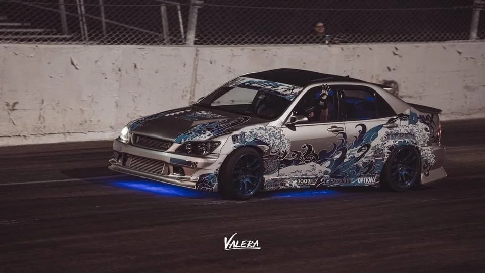 A modified racing car with a graphic wrap driving on a track at night, illuminated by blue underglow lights, and a car number decal on the side.