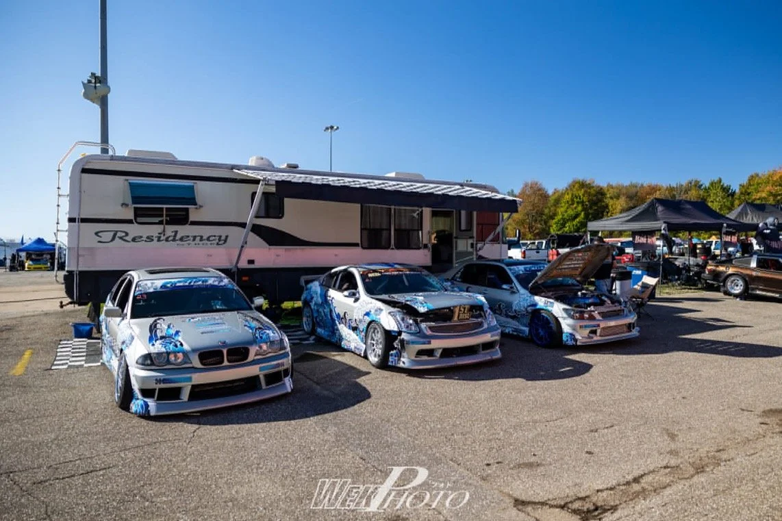 Three race cars parked in front of a large beige RV with an awning, at an outdoor motorsport event, with tents and other vehicles in the background on a sunny day.
